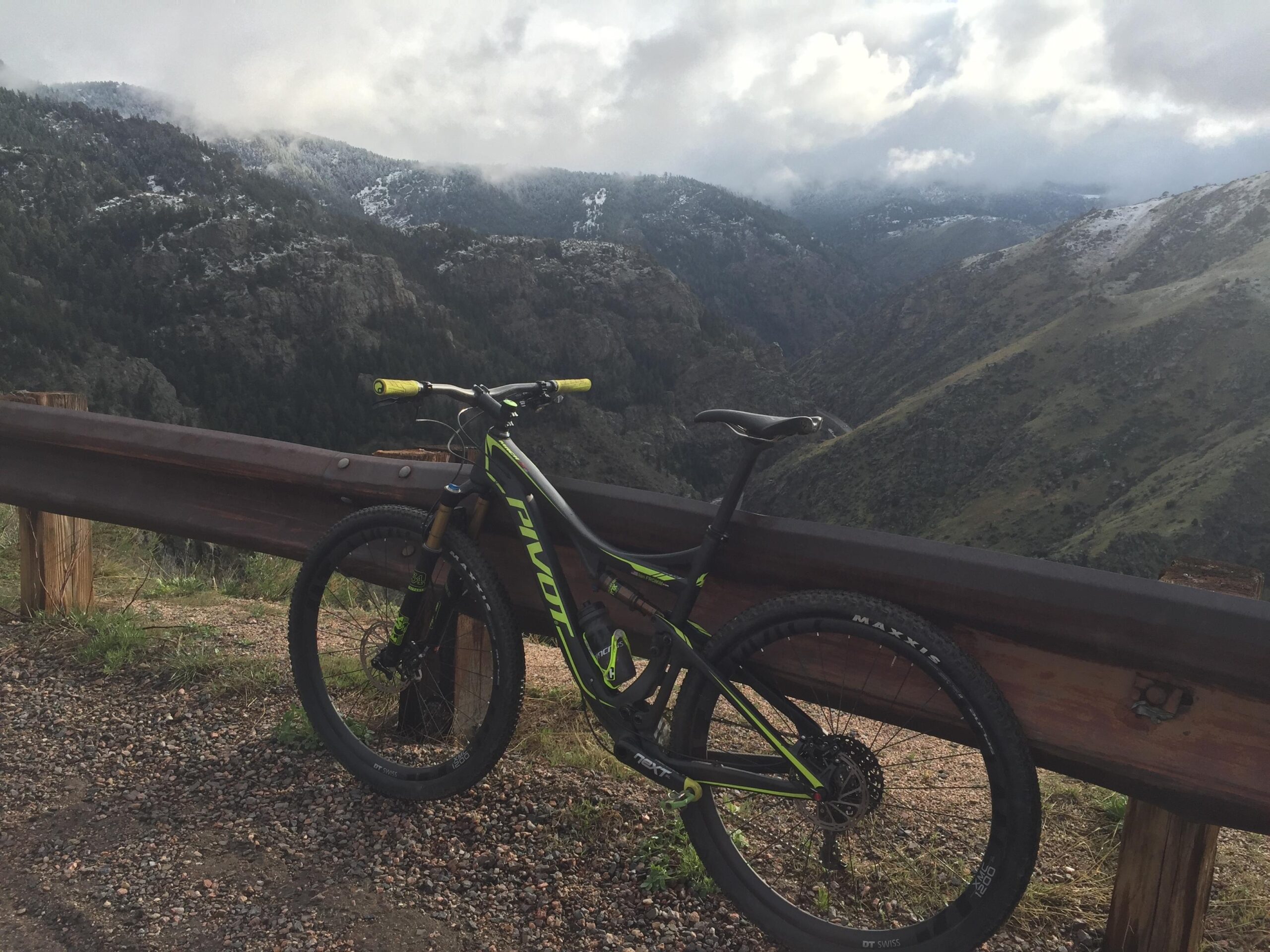 Pivot Mach 429SL Carbon: A mountain bike leaning against a guardrail with a scenic view of rocky hills and cloudy skies in the background. Some areas of the mountain are covered in snow, while the foreground features gravel and grass.