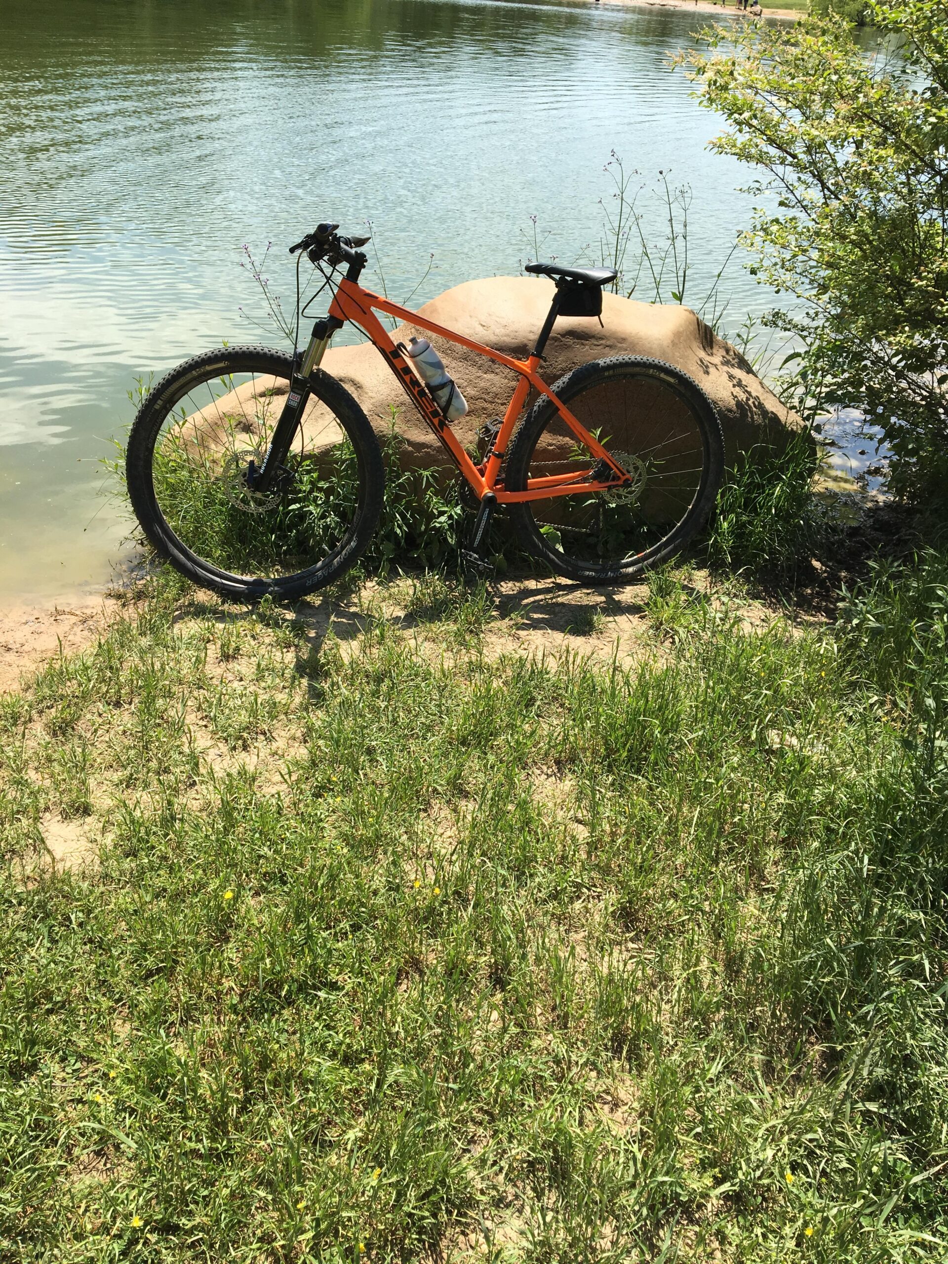 Trek Superfly 5: A bright orange mountain bike is leaned against a large rock by the edge of a calm, reflective lake. Surrounding the bike are patches of lush green grass and small wildflowers. The scene is sunny, with gentle ripples on the water's surface in the background.