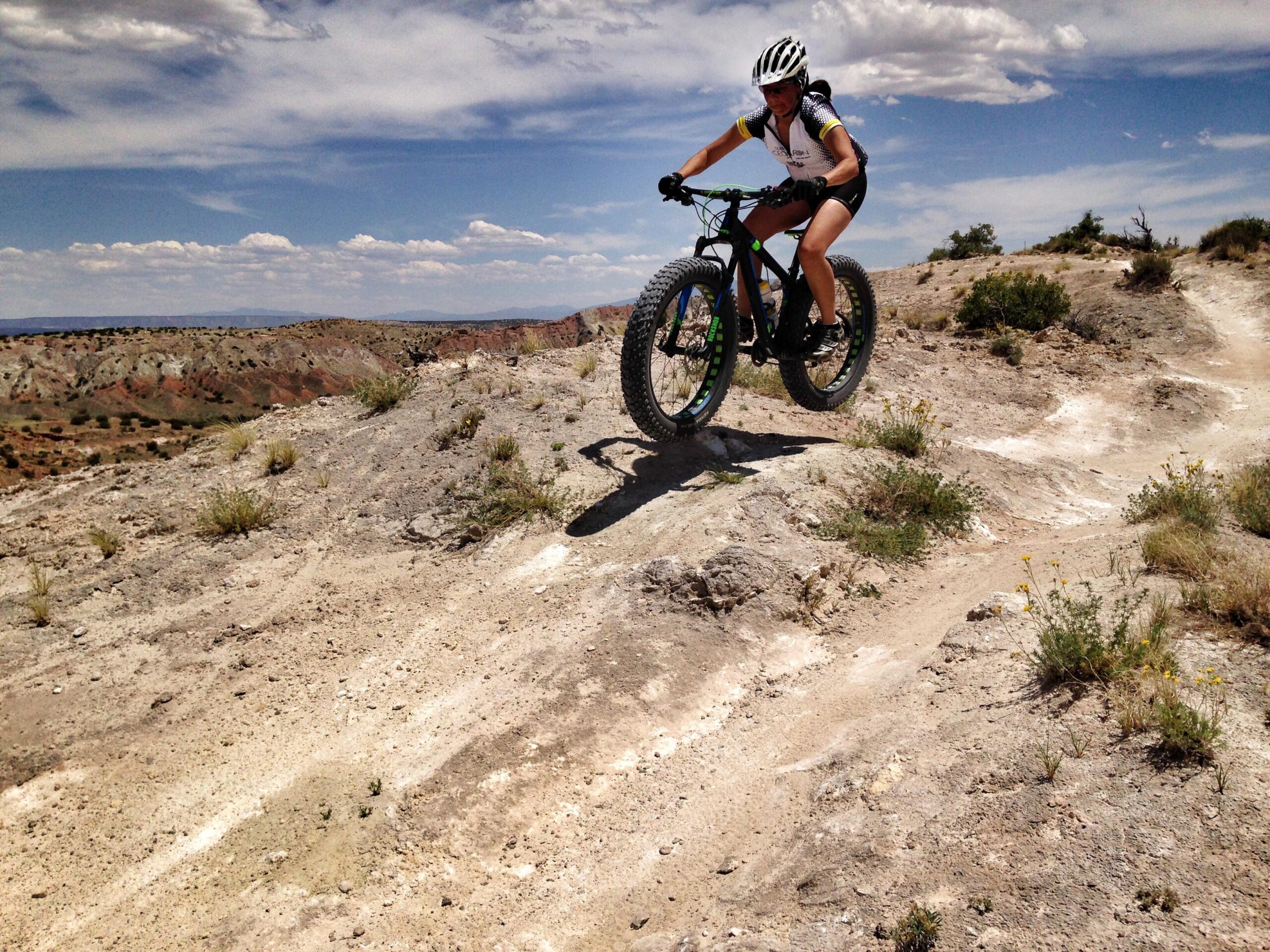 A cyclist riding a fat bike over rocky terrain, jumping off a small ledge with a scenic landscape of hills and cloudy skies in the background. White Ridge Bike Trails mountain bike trail.