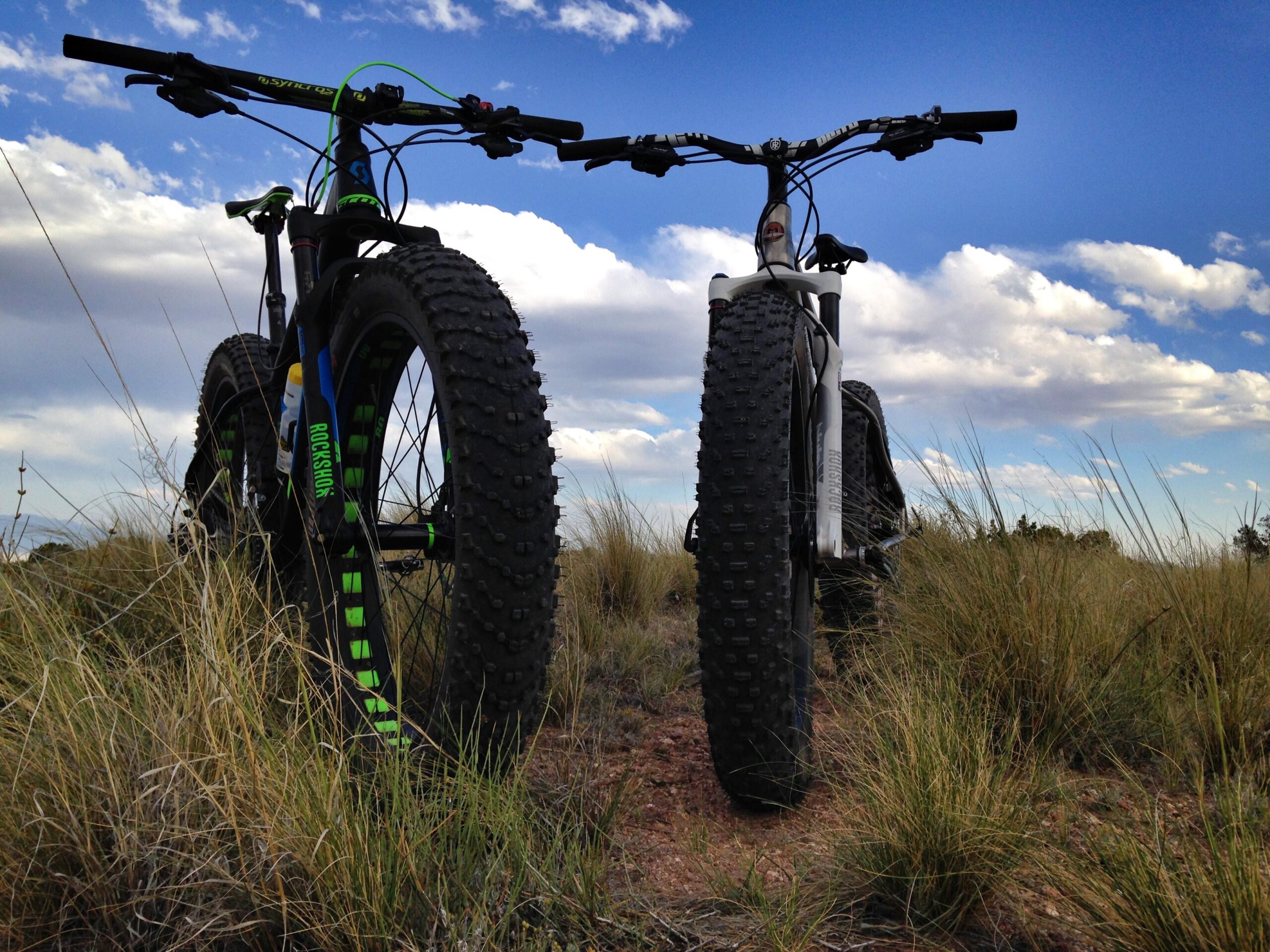 Two mountain bikes are positioned in a grassy field, with their rear tires facing the camera. The bikes have thick, knobby tires, and one is predominantly black with green accents, while the other is silver. Above the bikes, a blue sky is visible with scattered clouds. Mariposa Fat Bike Trails mountain bike trail.