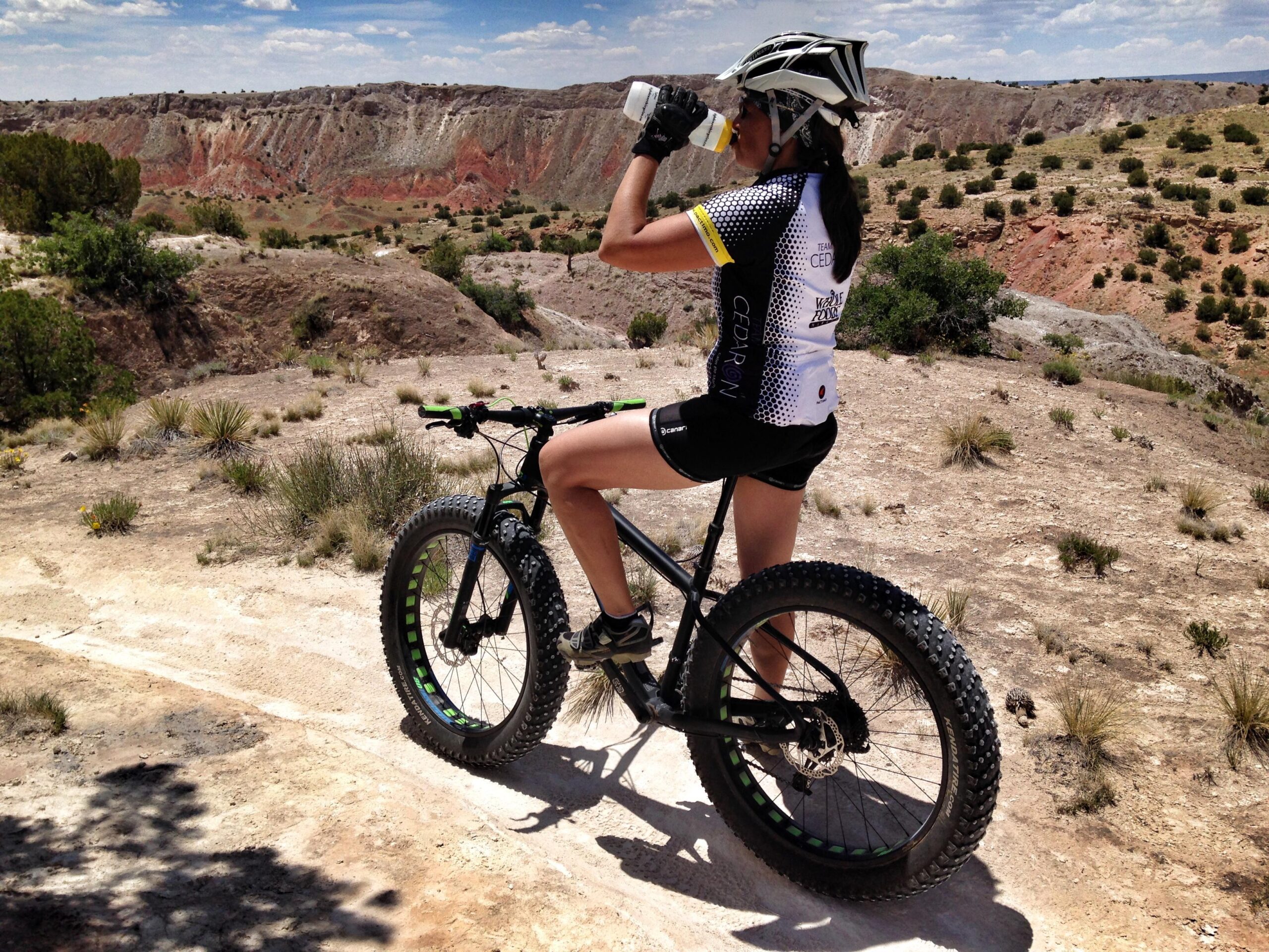 A woman wearing a cycling outfit, including a helmet, stops on a mountain bike to drink water while overlooking a rocky landscape with colorful hills and sparse vegetation. White Ridge Bike Trails mountain bike trail.