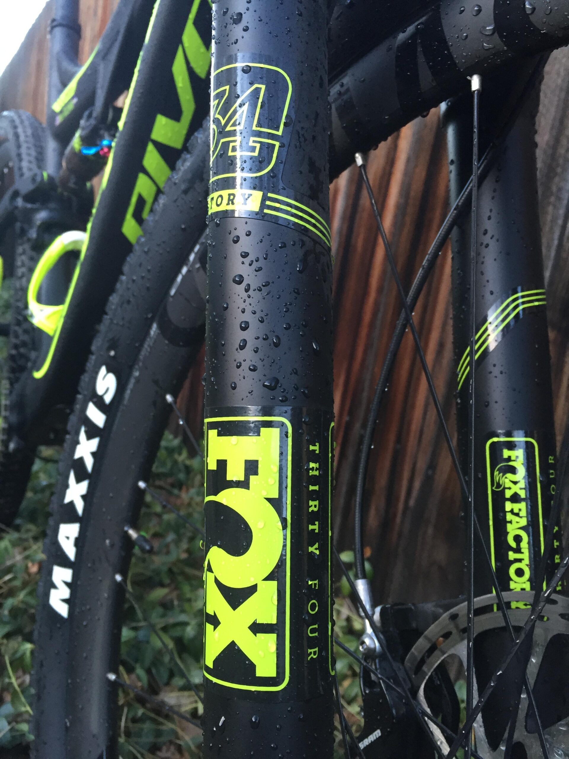 Pivot Mach 429SL Carbon: Close-up view of a mountain bike's frame with water droplets, showcasing black and neon green decals that read "FOX 34" and "FOX FACTORY." The bike features components from Maxxis. The background is blurred, highlighting the bike's design and graphics.