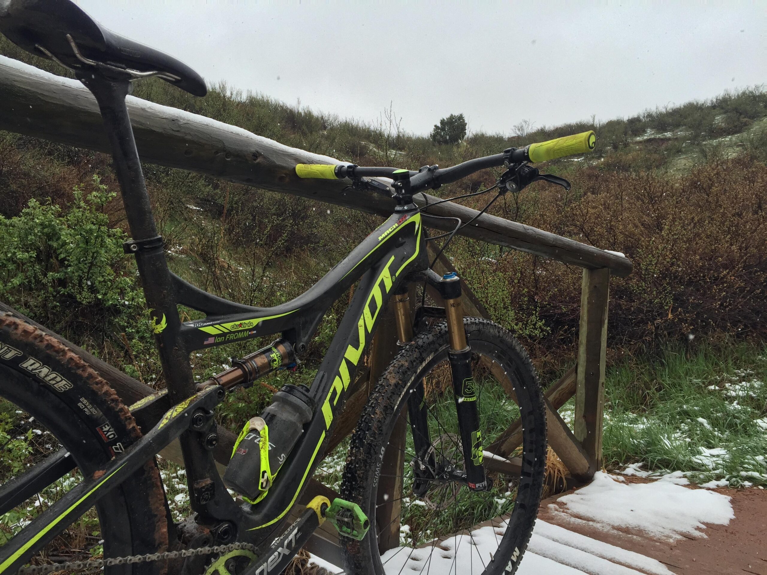 Pivot Mach 429SL Carbon: A close-up view of a mountain bike parked next to a wooden railing on a snowy trail. The bike features a black and green frame with a visible suspension system, dirt on the tires, and bright yellow grips. Snow covers parts of the ground and railing, while greenery is visible in the background under a cloudy sky.