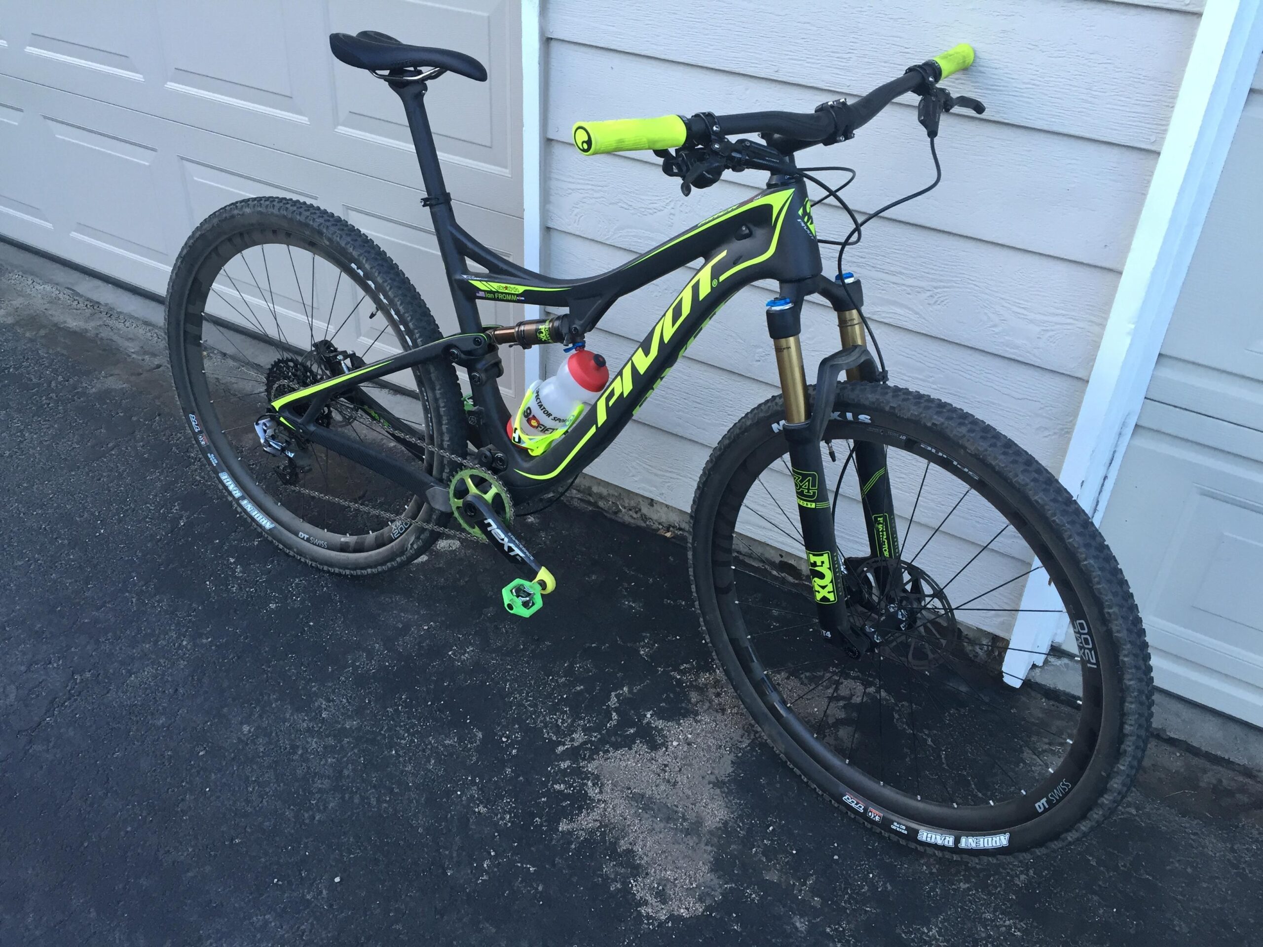 Pivot Mach 429SL Carbon: A black and neon green mountain bike is parked beside a garage door. The bike features thick tires, a water bottle holder, and bright green handlebars. The background shows a concrete surface and a portion of the garage wall.