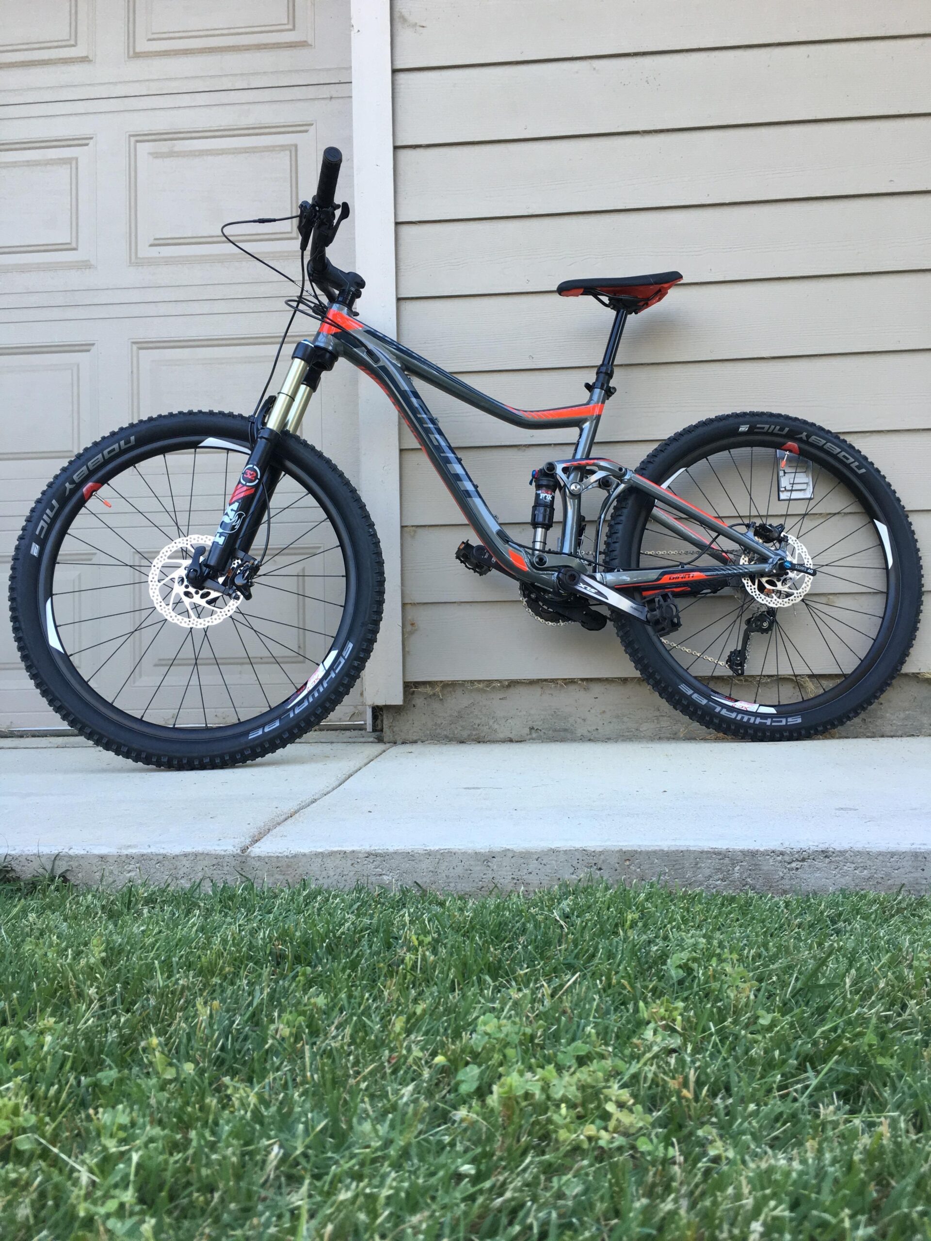 Giant Giant Trance 27.5 2: A mountain bike with a gray and orange frame, featuring large tires, disc brakes, and a modern suspension system, positioned against a light-colored wooden wall with a nearby patch of green grass.