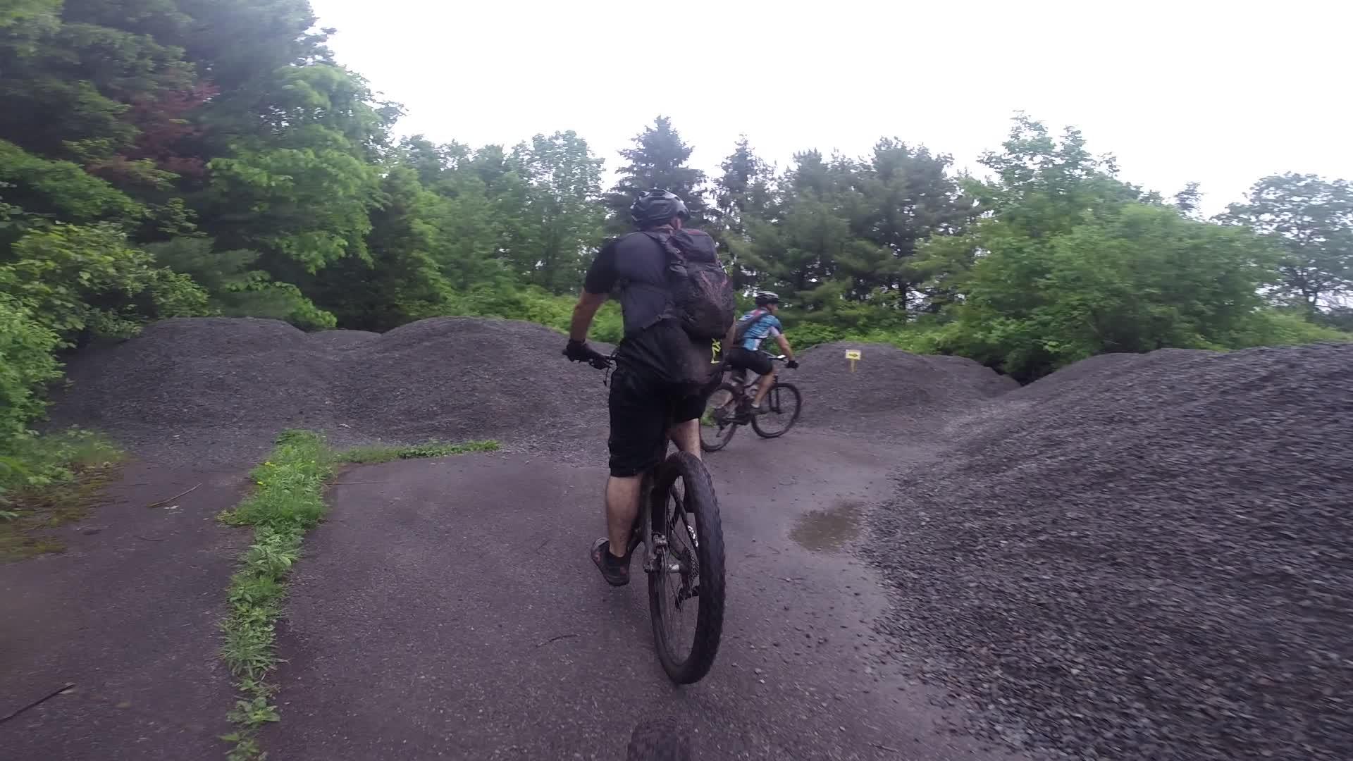 Two mountain bikers ride on a gravel path surrounded by greenery and dirt mounds. The riders are wearing helmets and riding gear, focused on navigating the terrain. The atmosphere appears adventurous, with a cloudy sky overhead. Stewart State Forest mountain bike trail.