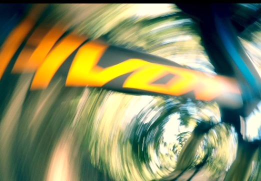 Pivot Mach 6 Carbon: An abstract image featuring a blurred orange logo against a backdrop of swirling green and brown foliage, suggesting motion and speed. The focus is on the logo, which appears dynamic amidst the surrounding colors and shapes.