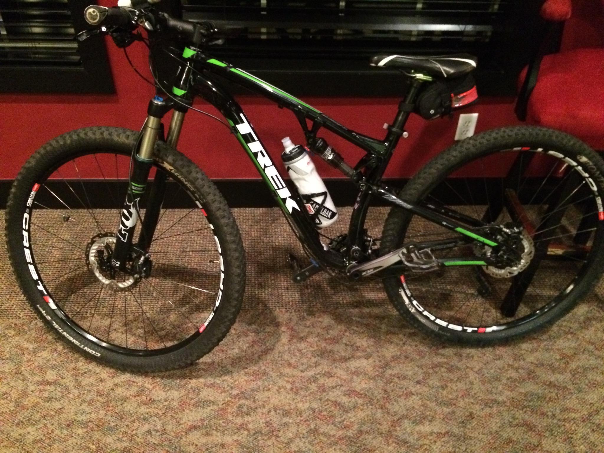 Trek Superfly FS 8: A black and green Trek mountain bike resting on a carpeted floor. The bike features thick tires, front suspension, and a water bottle attached to the frame. In the background, there is a red chair and partly visible furnishings.
