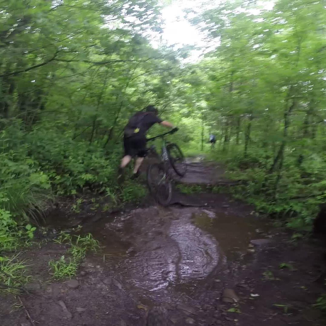 A mountain biker jumping over a muddy patch on a forest trail, surrounded by lush green foliage. The rider is focused on maneuvering through the challenging terrain, with another cyclist visible in the background. Stewart State Forest mountain bike trail.