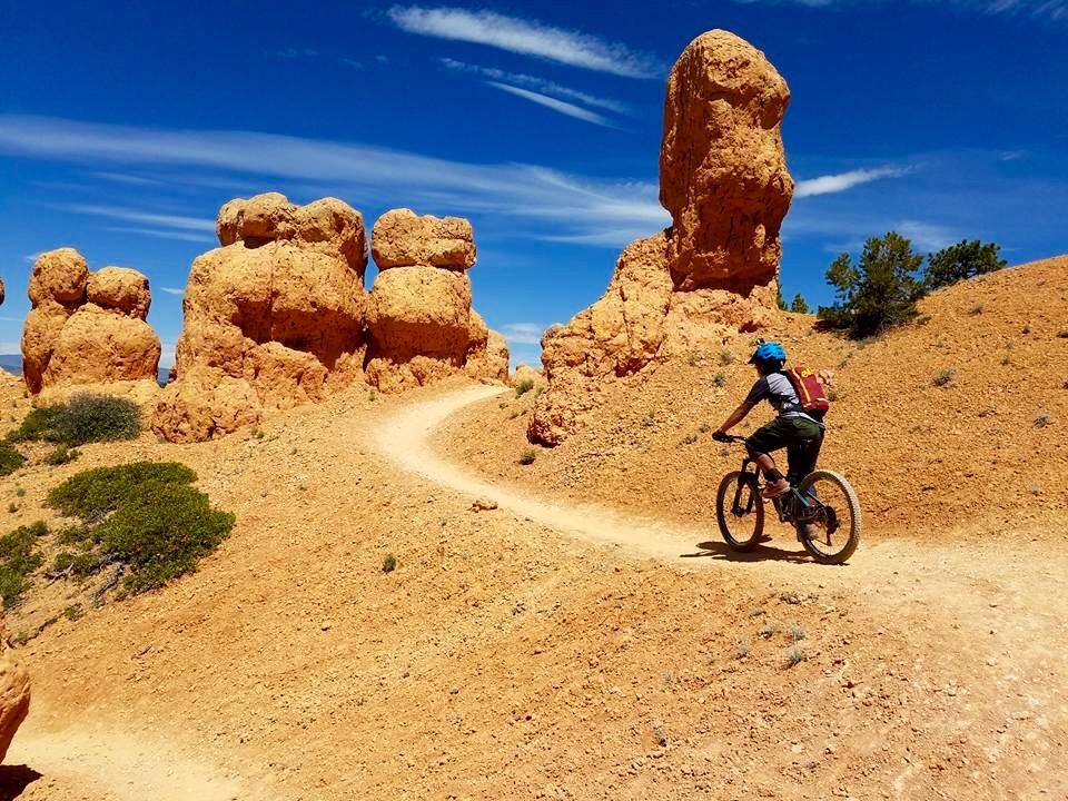 A person rides a mountain bike along a dusty trail surrounded by unique rock formations under a clear blue sky. The terrain is arid, featuring orange-hued earth and scattered greenery. Thunder Mountain mountain bike trail.