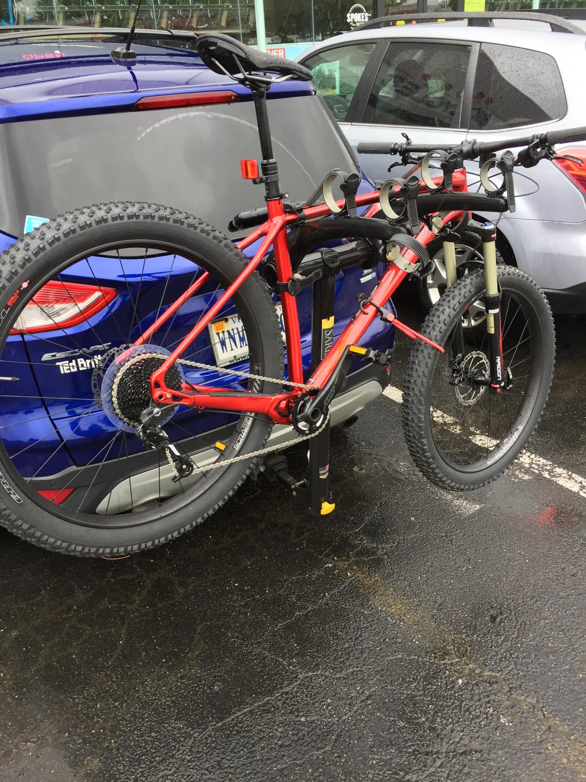 Specialized Fuse Comp 6Fattie: A bright red mountain bike mounted on a bike rack attached to the back of a blue vehicle in a parking lot, with wet pavement visible from recent rain. The bike features thick tires and a visible gear system, while the vehicle displays a license plate and some stickers.