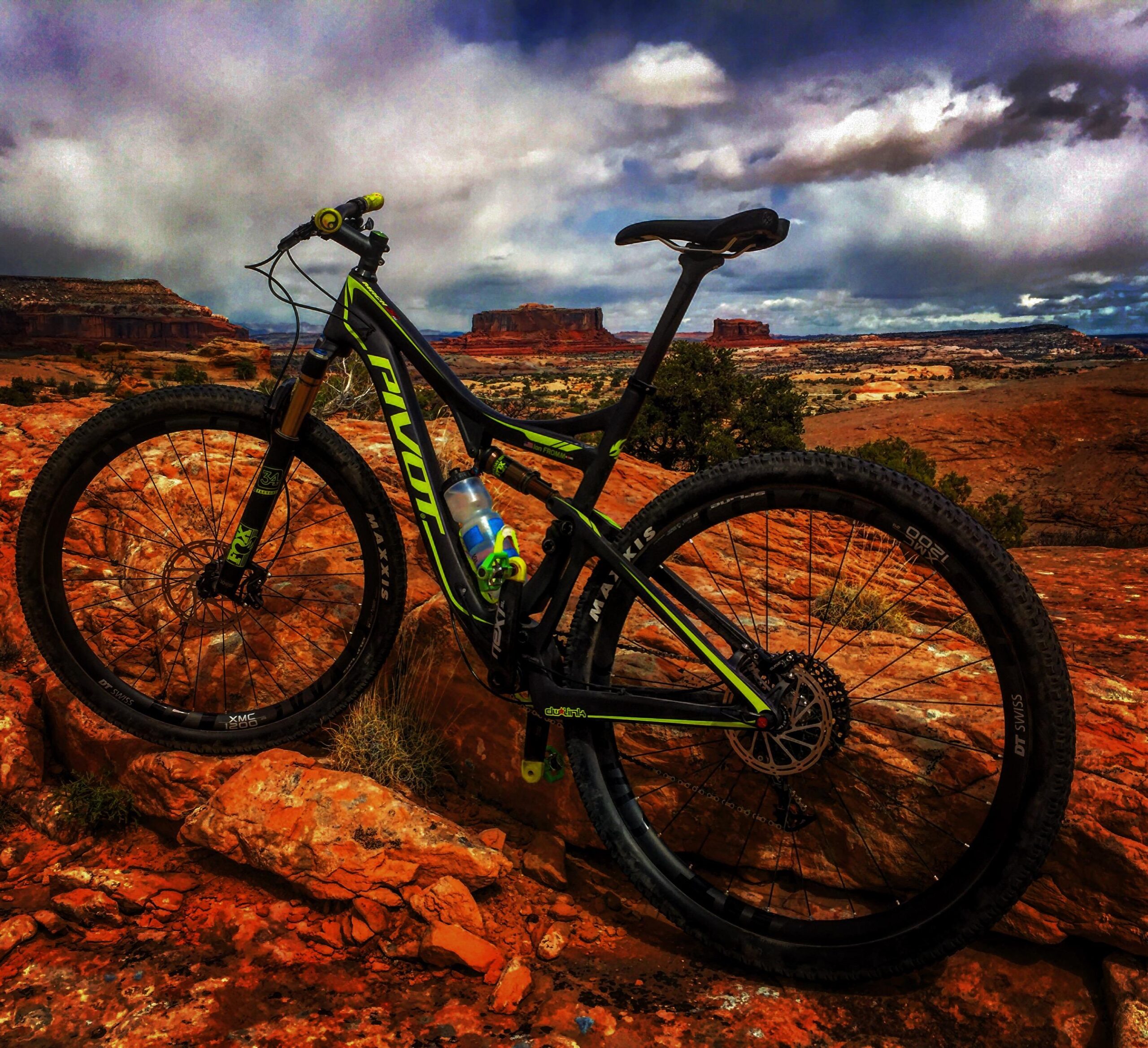 Pivot Mach 429SL Carbon: A mountain bike parked on rocky terrain with a scenic backdrop of red cliffs and a cloudy sky. The bike features a black and green design and has a water bottle mounted on the frame.