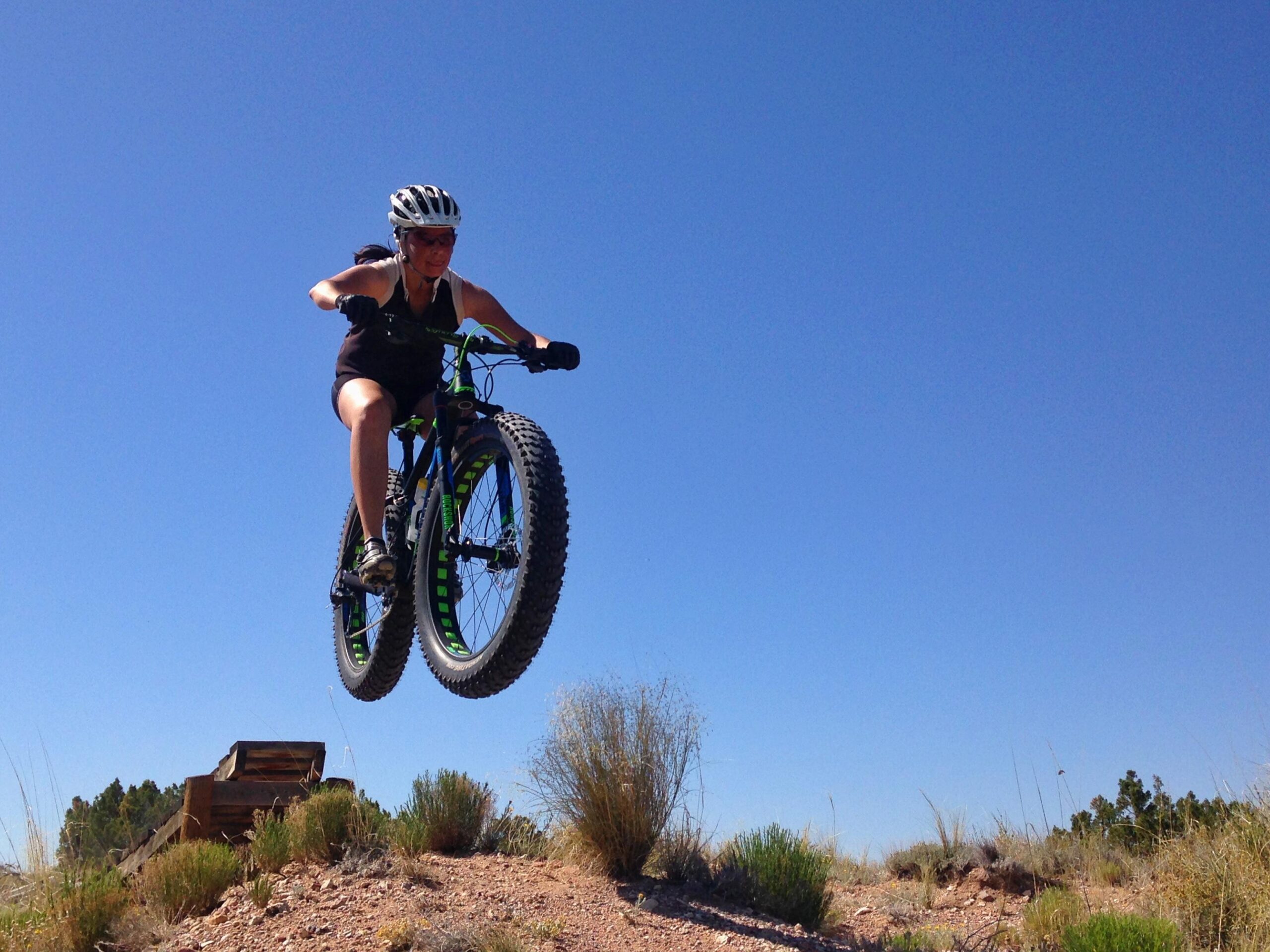 A person in a black tank top and shorts is riding a mountain bike, airborne as they jump off a small dirt ramp. The sky is clear and blue, with shrubs and grasses visible in the foreground and background. The bike has large tires and vibrant green accents. Mariposa Fat Bike Trails mountain bike trail.