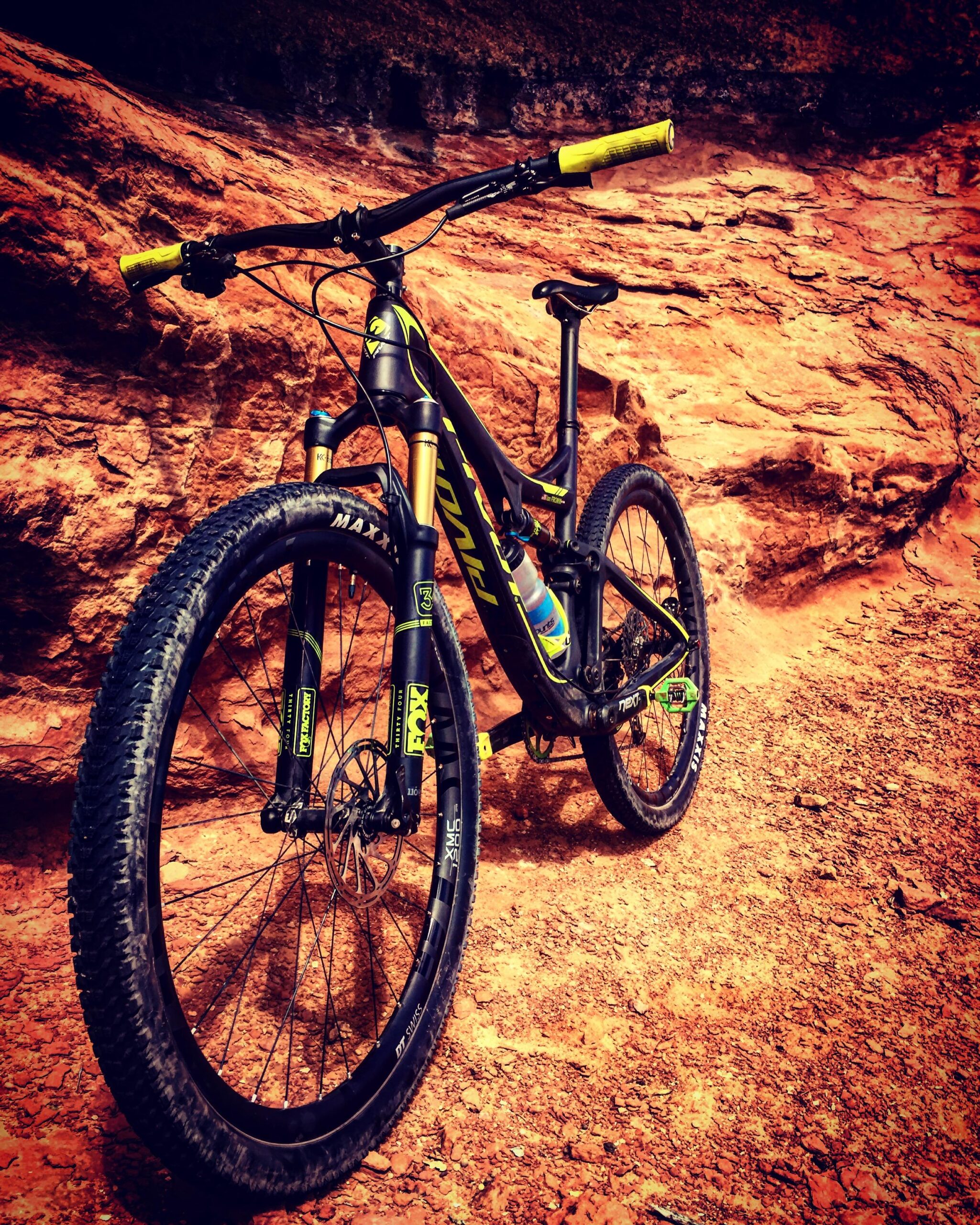 Pivot Mach 429SL Carbon: A mountain bike positioned on a rocky terrain, showcasing its black and yellow frame, thick tires, and prominent suspension forks. The background features rugged red rock formations, emphasizing an outdoor adventure setting.