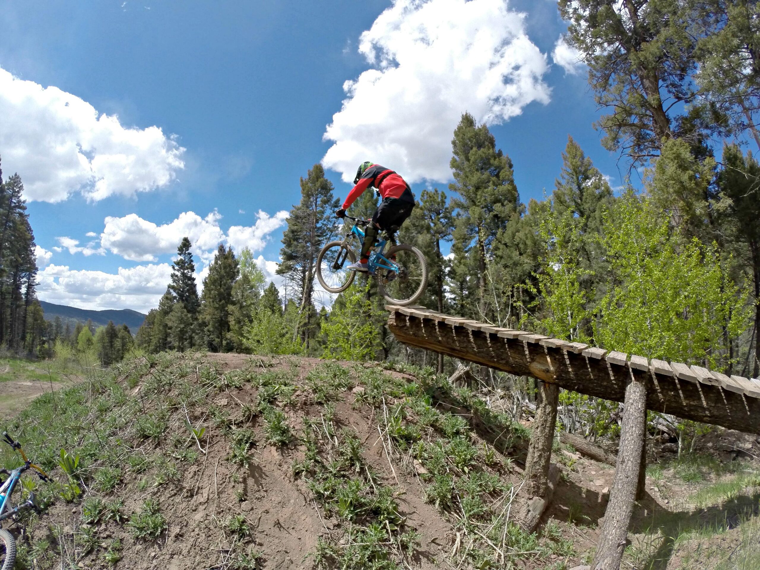 A mountain biker in a red jersey jumps off a wooden ramp over a dirt mound in a forested area. The sky is partly cloudy, with blue patches visible, and greenery surrounds the scene, including trees and shrubs. Angel Fire Bike Park mountain bike trail.