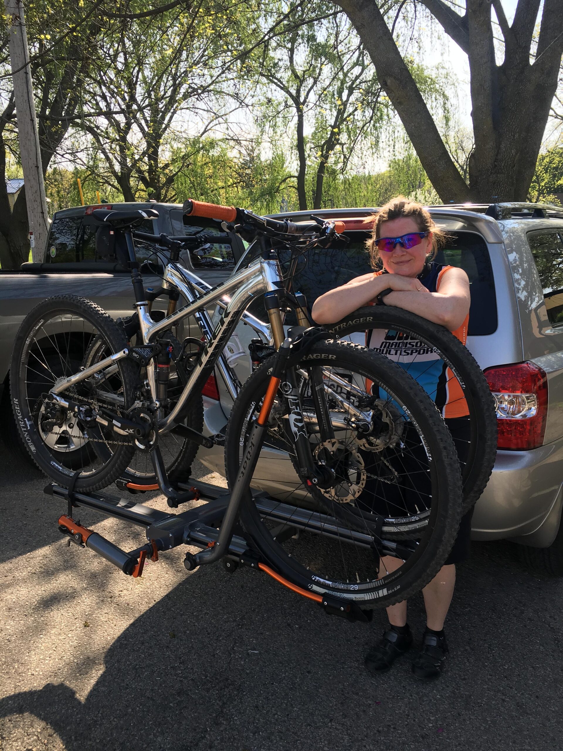Trek Fuel EX 9 29: A person in an orange and blue cycling outfit leans against a car while holding onto a mountain bike. The bike is mounted on a bike rack attached to the rear of the vehicle. The background features trees and a sunny, clear sky.