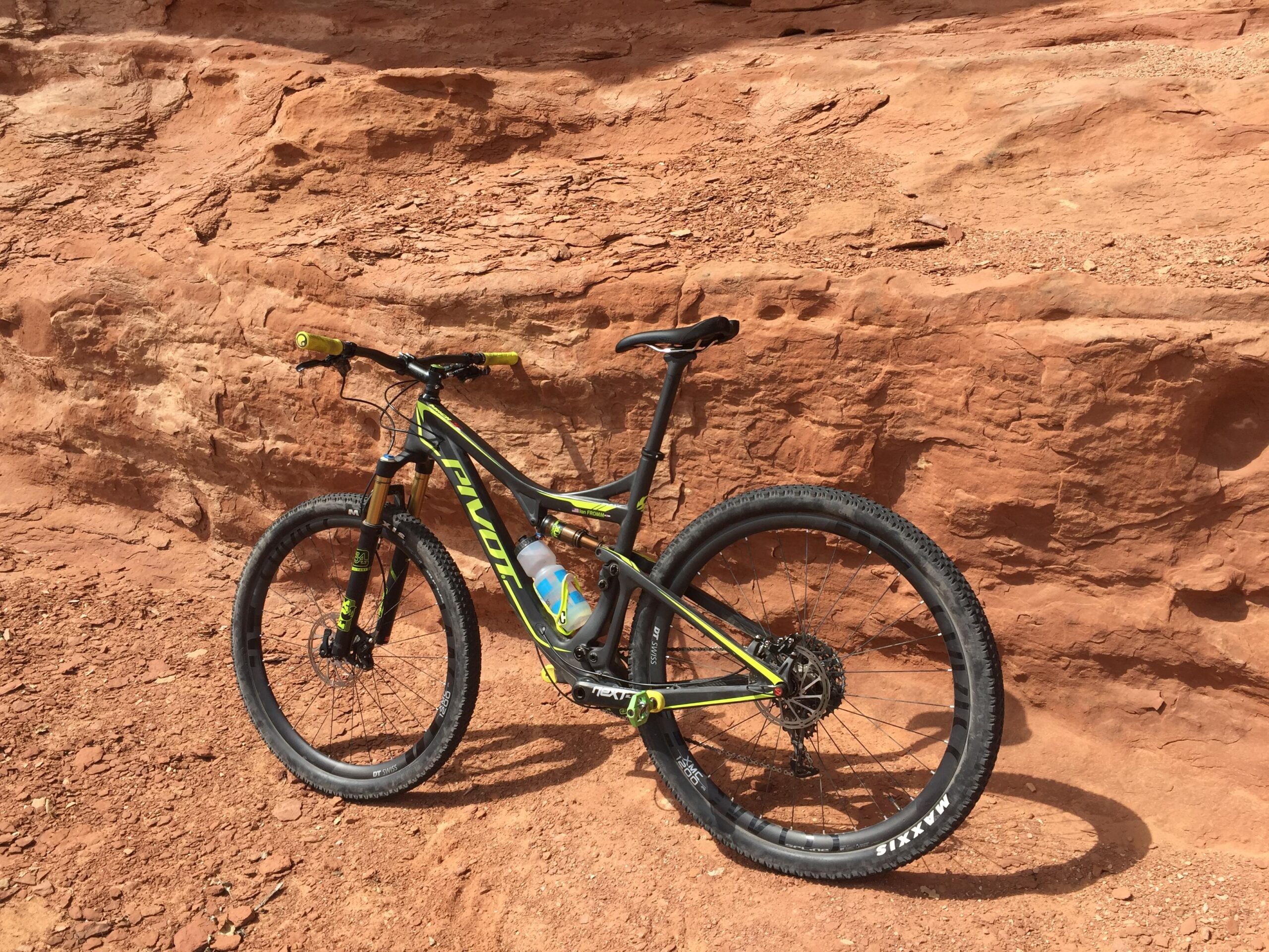 Pivot Mach 429SL Carbon: A mountain bike with a black and yellow frame, positioned on a rocky, reddish terrain. The bike features thick tires suitable for off-road riding and a water bottle attached to the frame, with a rugged rock formation in the background.