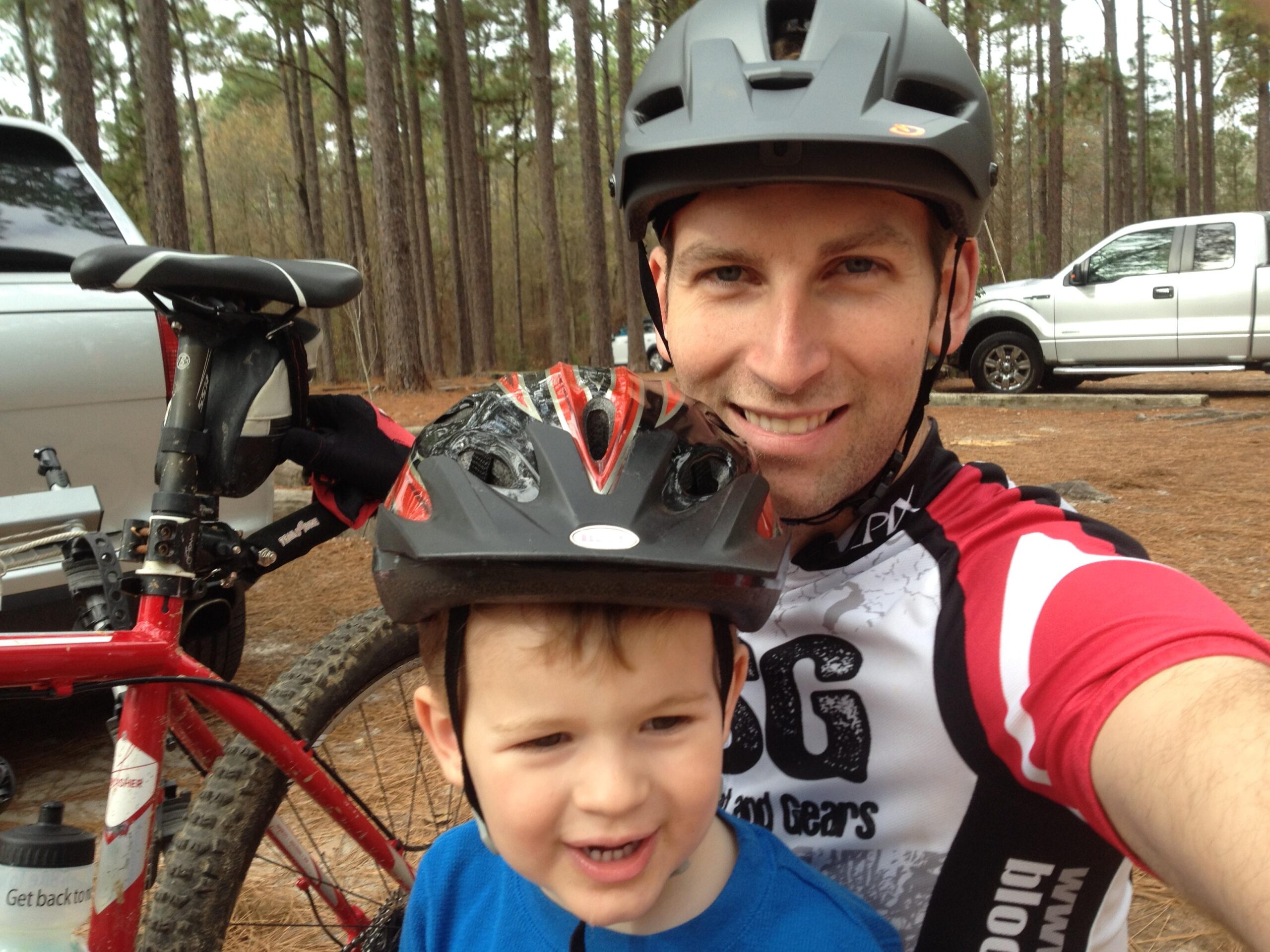 Trek 2016 Trek Top Fuel 8: A man and a young boy are posing for a selfie in a wooded area, both wearing helmets. The man is smiling and dressed in a cycling jersey, while the boy, in a blue shirt, is also smiling. In the background, there is a red mountain bike and a parked vehicle. The scene conveys a cheerful and active outdoor moment.