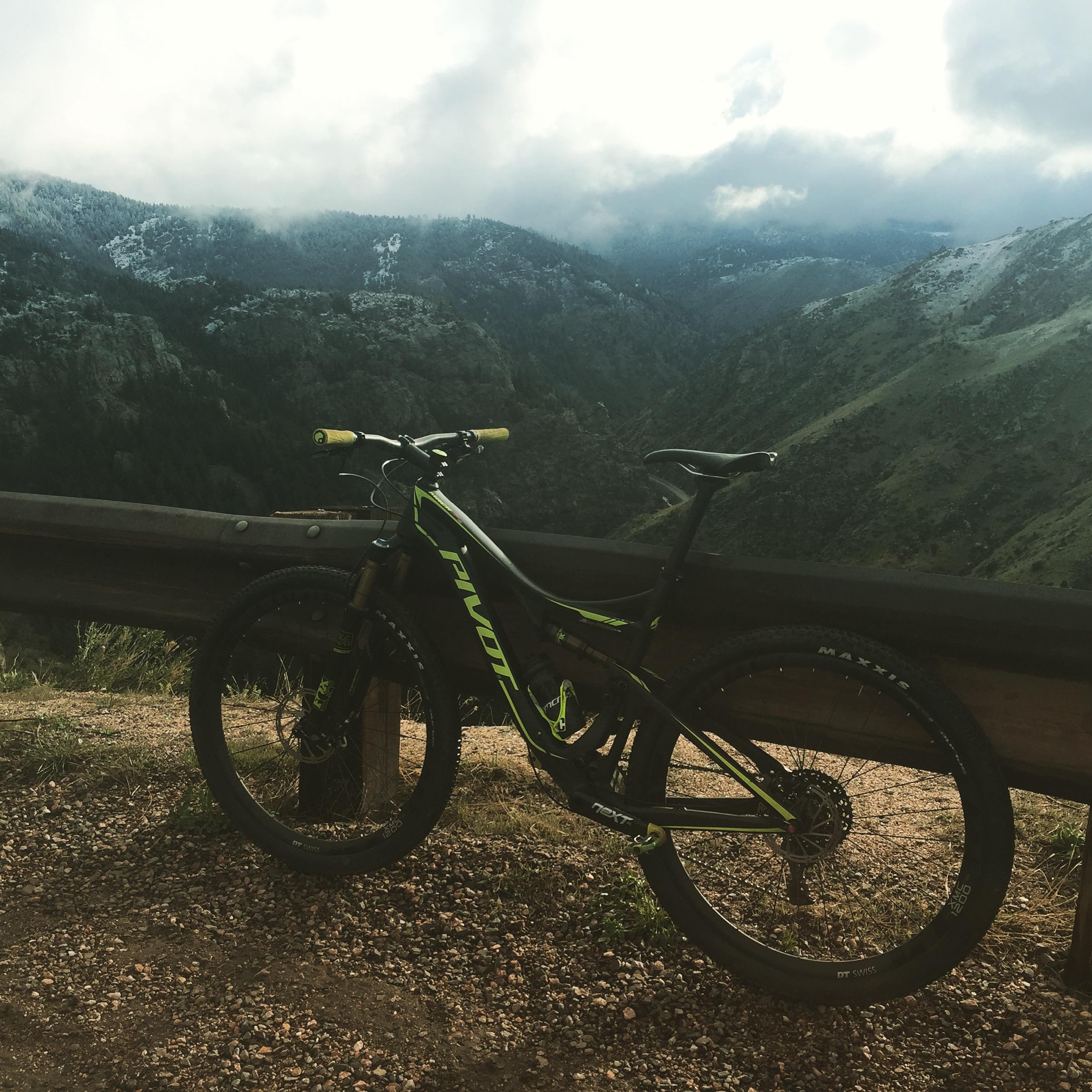 Pivot Mach 429SL Carbon: A mountain bike parked next to a wooden railing, overlooking a scenic mountainous landscape with hills and valleys under a cloudy sky. The bike features black and bright green colors, sitting on gravel terrain.