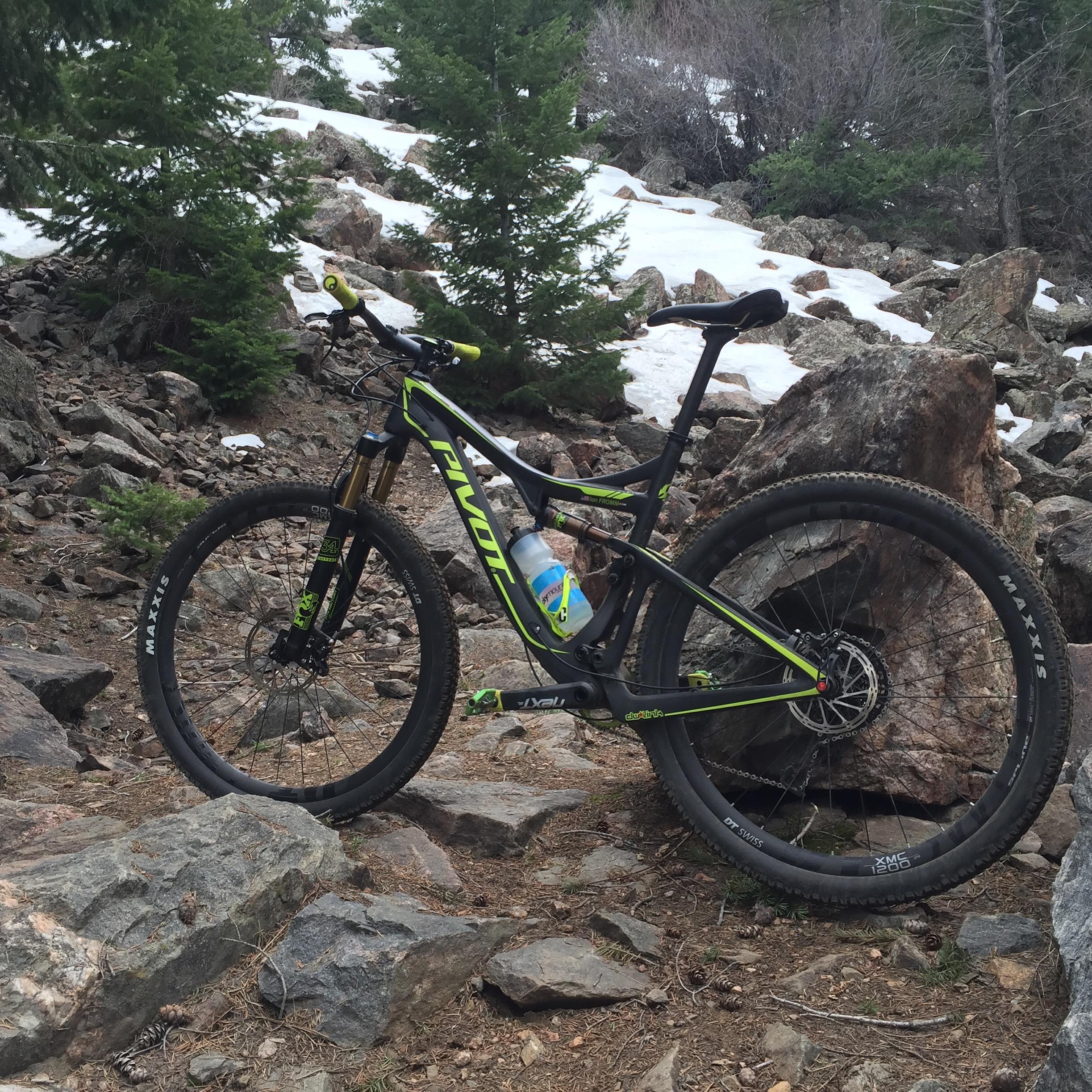 Pivot Mach 429SL Carbon: A mountain bike parked on rocky terrain, surrounded by pine trees and patches of snow, with a water bottle mounted on the frame.