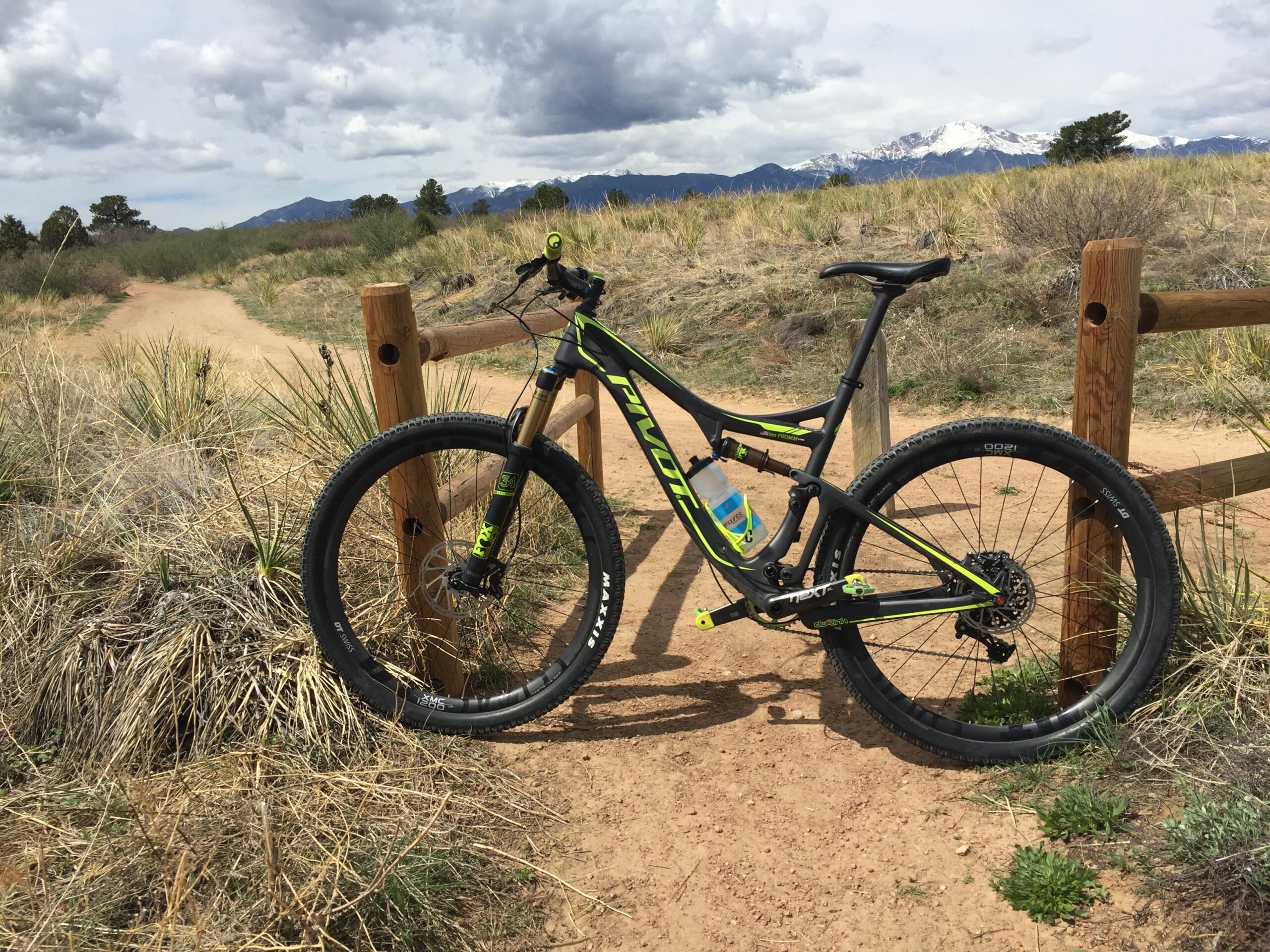 Pivot Mach 429SL Carbon: A mountain bike resting against a wooden fence on a dirt trail, surrounded by tall grass and shrubs. In the background, there are mountains with patches of snow under a cloudy sky.