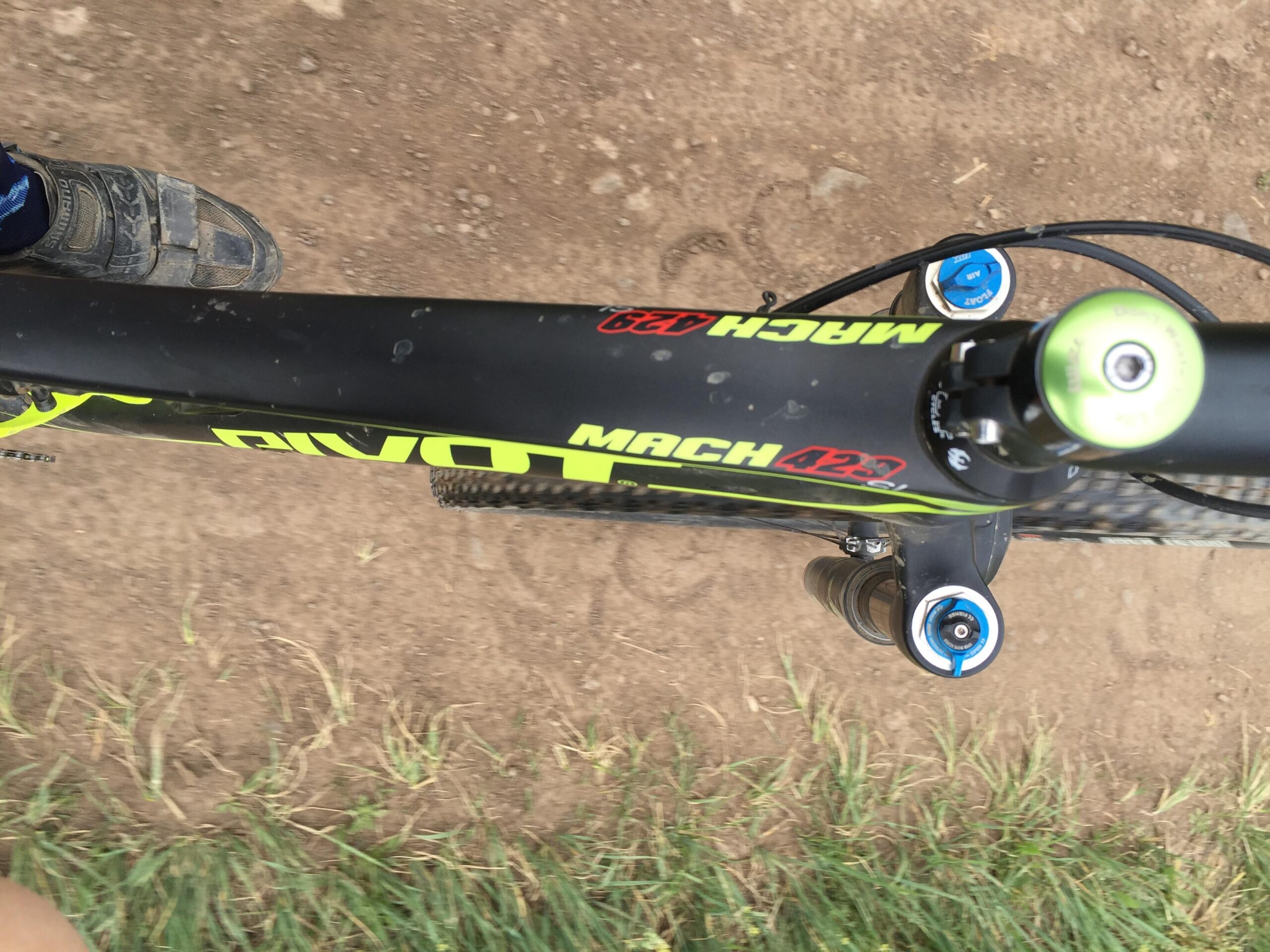 Pivot Mach 429SL Carbon: A top-down view of a mountain bike's frame and handlebars, featuring the "Pivot Mach 429" logo prominently on the crossbar. The image includes a rider's foot on the pedal and a patch of dirt and grass below, indicating an outdoor biking environment.