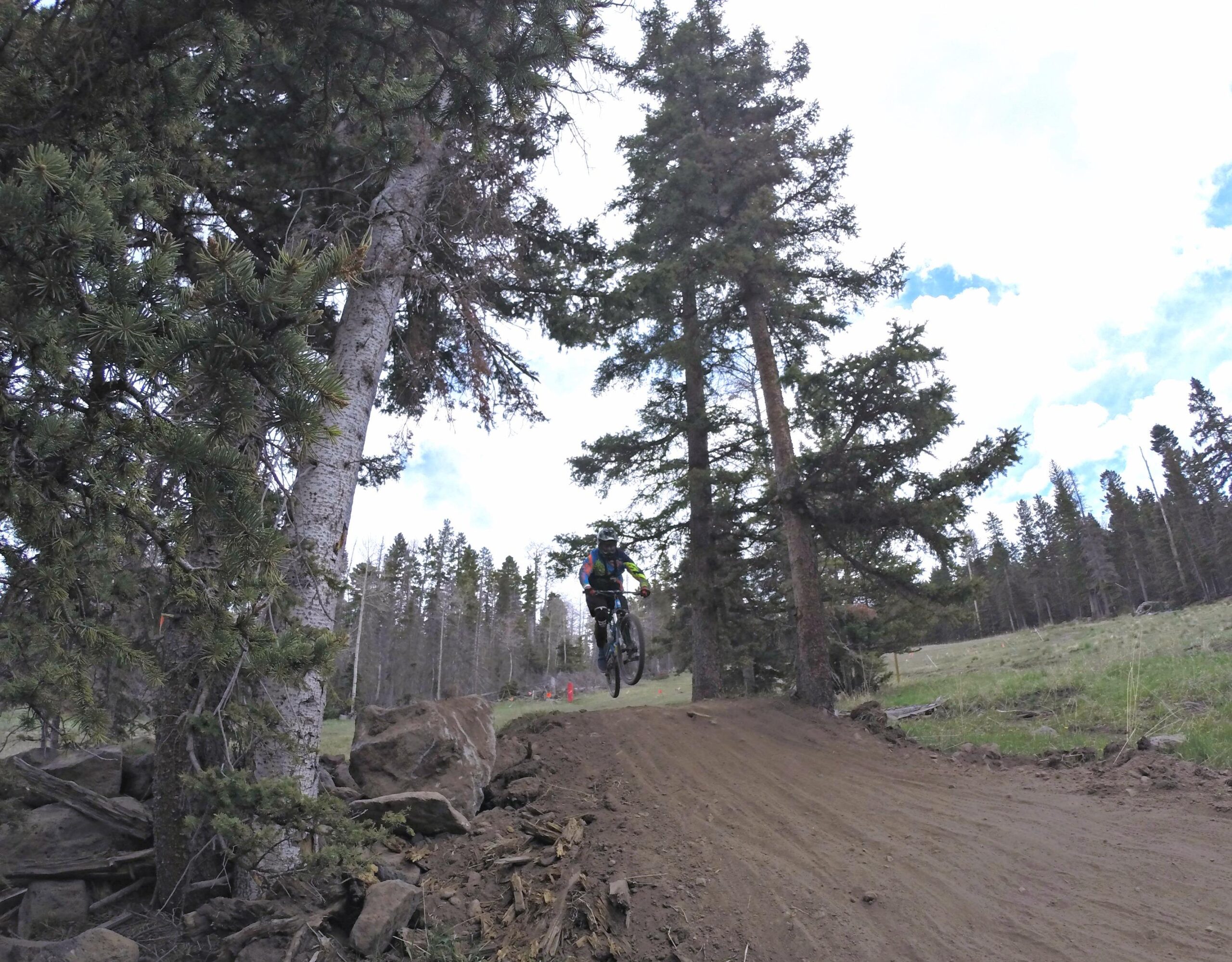 A mountain biker jumping off a dirt ramp surrounded by tall trees in a forested area. The sky is partly cloudy, and the landscape is green and natural, showcasing a vibrant outdoor setting for biking. Angel Fire Bike Park mountain bike trail.