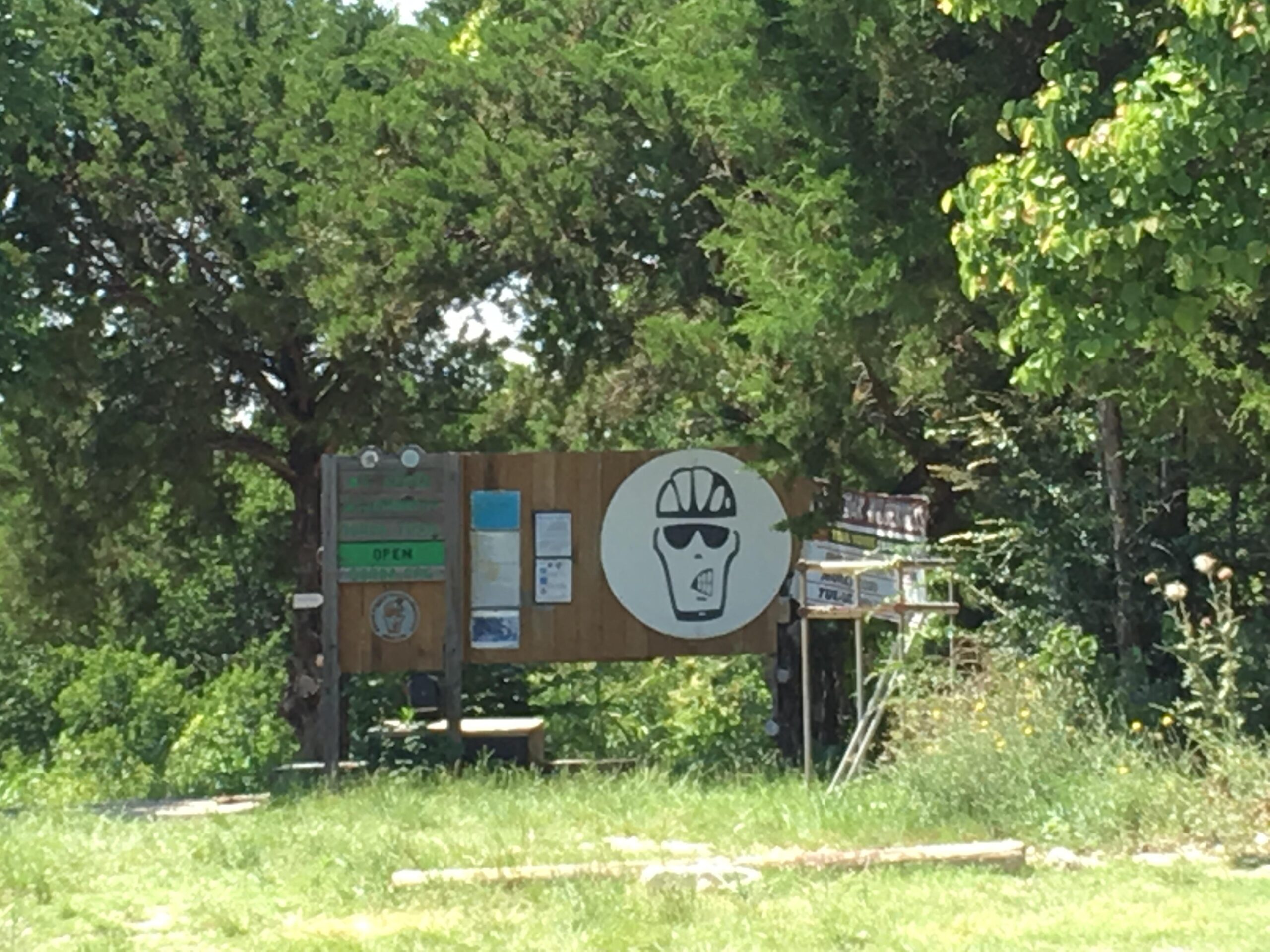 Signage at a park entrance featuring a large, stylized graphic of a person wearing a helmet and sunglasses. The sign includes a green "OPEN" indication and various posted notices. Surrounding the sign are trees and tall grass, suggesting a natural outdoor environment. Big Cedar Wilderness Trails mountain bike trail.