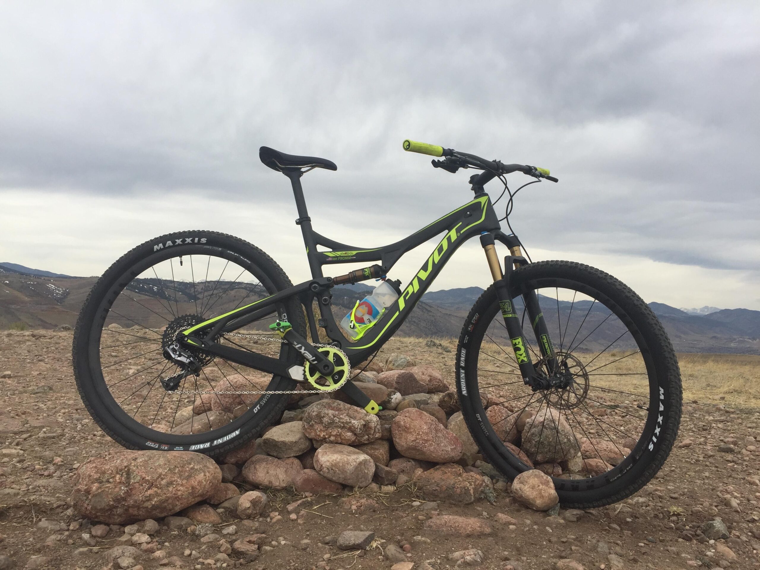 Pivot Mach 429SL Carbon: A mountain bike leaning against a small rock pile, set against a cloudy sky and rolling hills in the background. The bike features a black and bright green frame, Maxxis tires, and a colorful pedal.