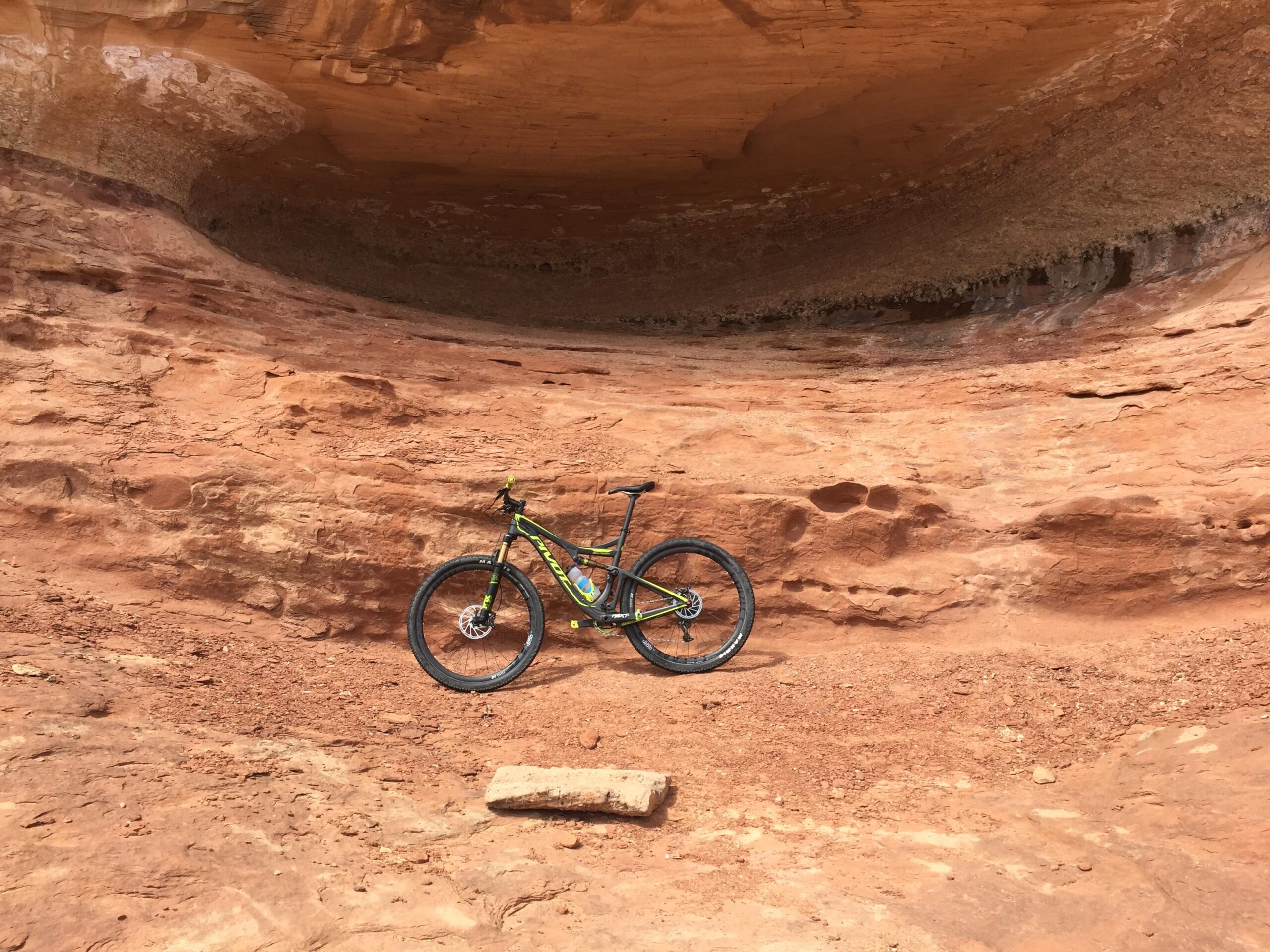Pivot Mach 429SL Carbon: A mountain bike rests against a rocky, reddish-brown terrain beneath a large, overhanging rock formation. The surrounding environment showcases natural erosion patterns in the sandstone, creating a rugged outdoor setting.