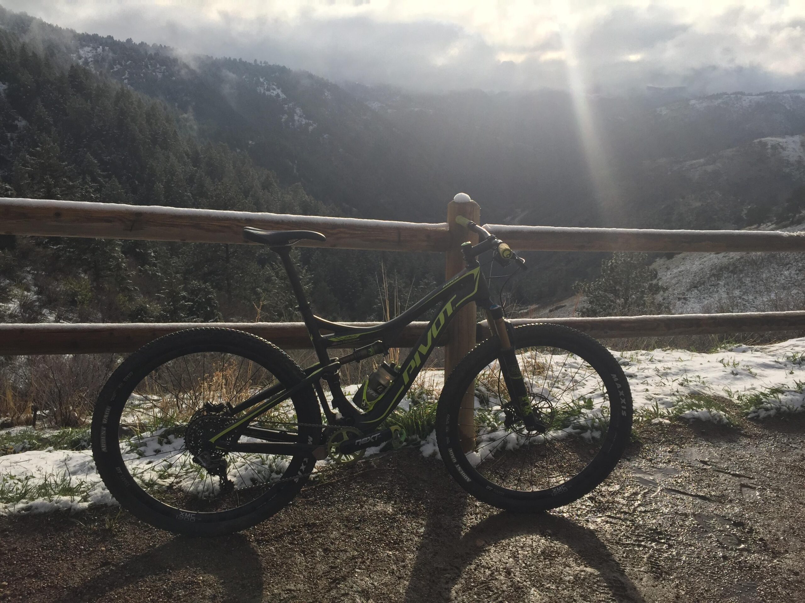 Pivot Mach 429SL Carbon: A mountain bike leaning against a wooden fence with a scenic view of snow-dusted mountains and greenery in the background. The sun is shining through the clouds, creating a dramatic lighting effect.