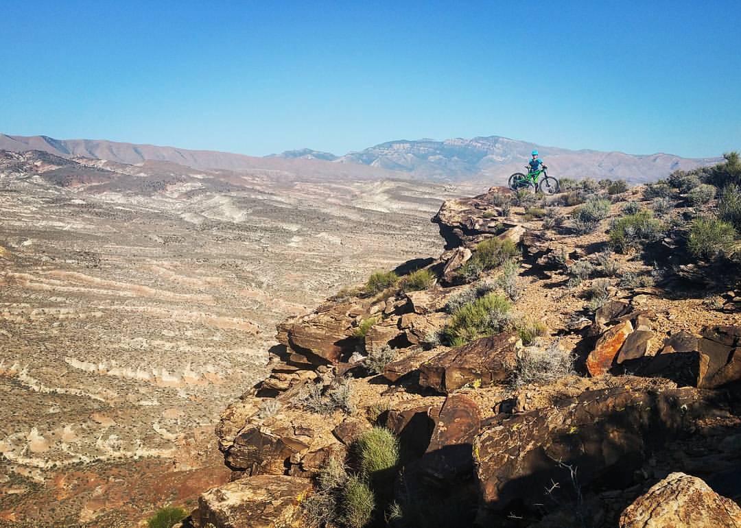 A mountain biker stands on the edge of a rocky cliff, overlooking a vast desert landscape with rolling hills and layered rock formations under a clear blue sky. Sparse vegetation grows among the rocks, highlighting the rugged terrain. Barrel Roll mountain bike trail.