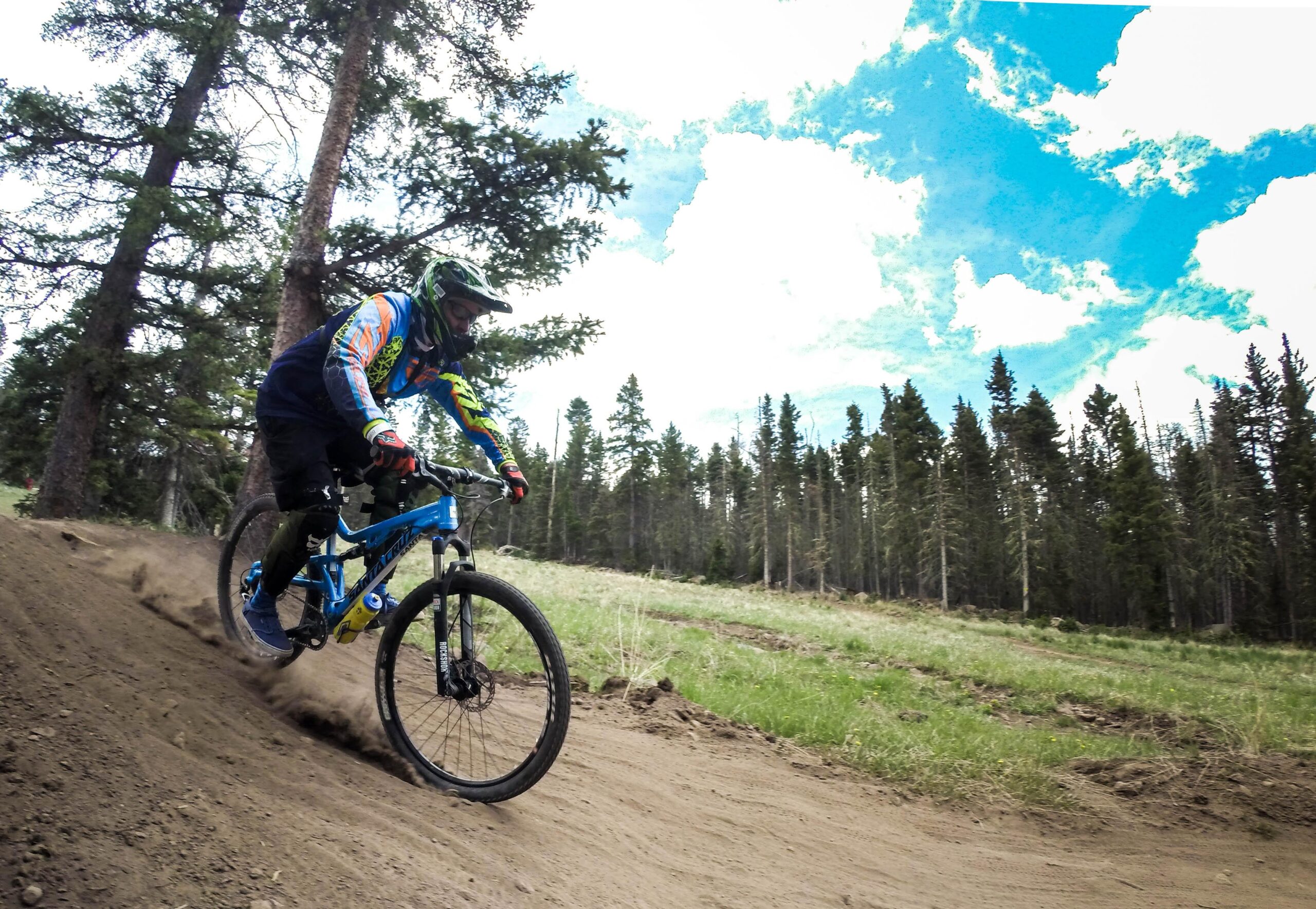A mountain biker riding through a dirt trail in a forested area, with trees in the background and a blue sky with clouds above. The rider is wearing a helmet and colorful cycling gear, leaning into a turn as dust kicks up from the trail. Angel Fire Bike Park mountain bike trail.