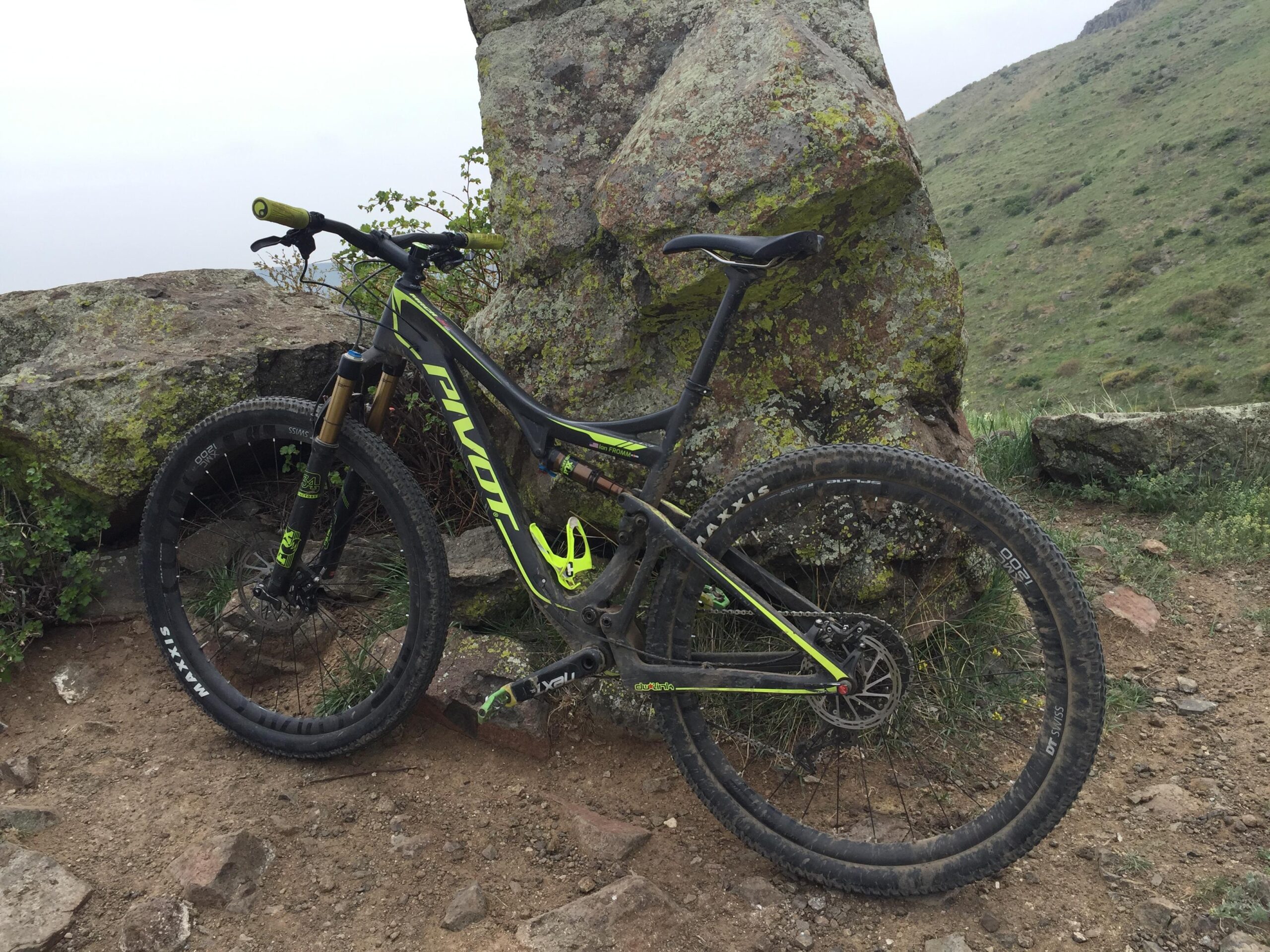 Pivot Mach 429SL Carbon: A black and green mountain bike is resting against a large, moss-covered rock, positioned on a dirt trail. The background features a lush, grassy hillside under a cloudy sky. The bike is equipped with sturdy tires and visible suspension components, indicating it's designed for rugged terrain.
