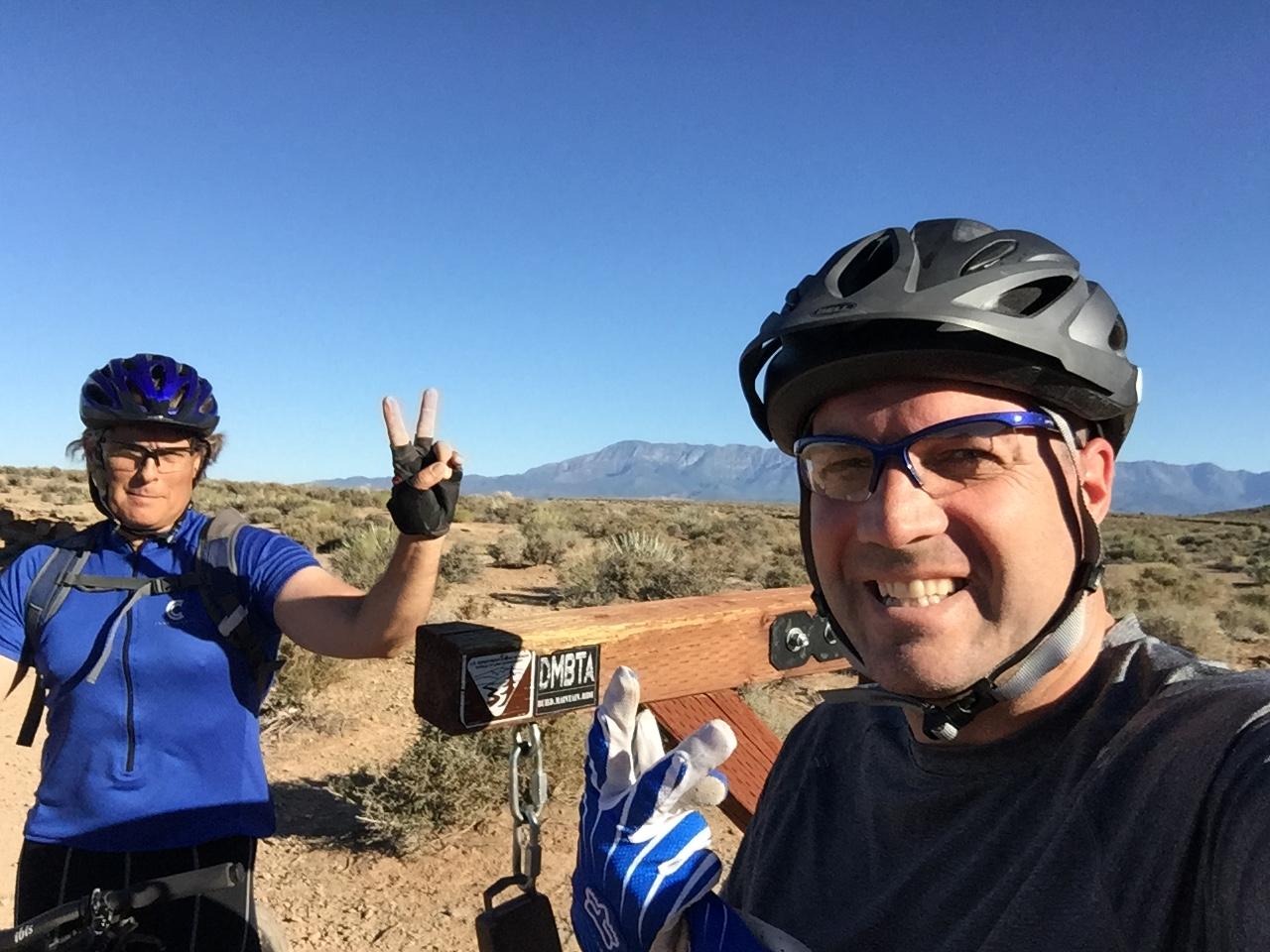 Two cyclists wearing helmets and gloves pose for a selfie in a desert landscape. One cyclist raises two fingers in a peace sign, while the other smiles and shows a thumbs-up. The background features mountains and clear blue skies. J.E.M. Trail mountain bike trail.