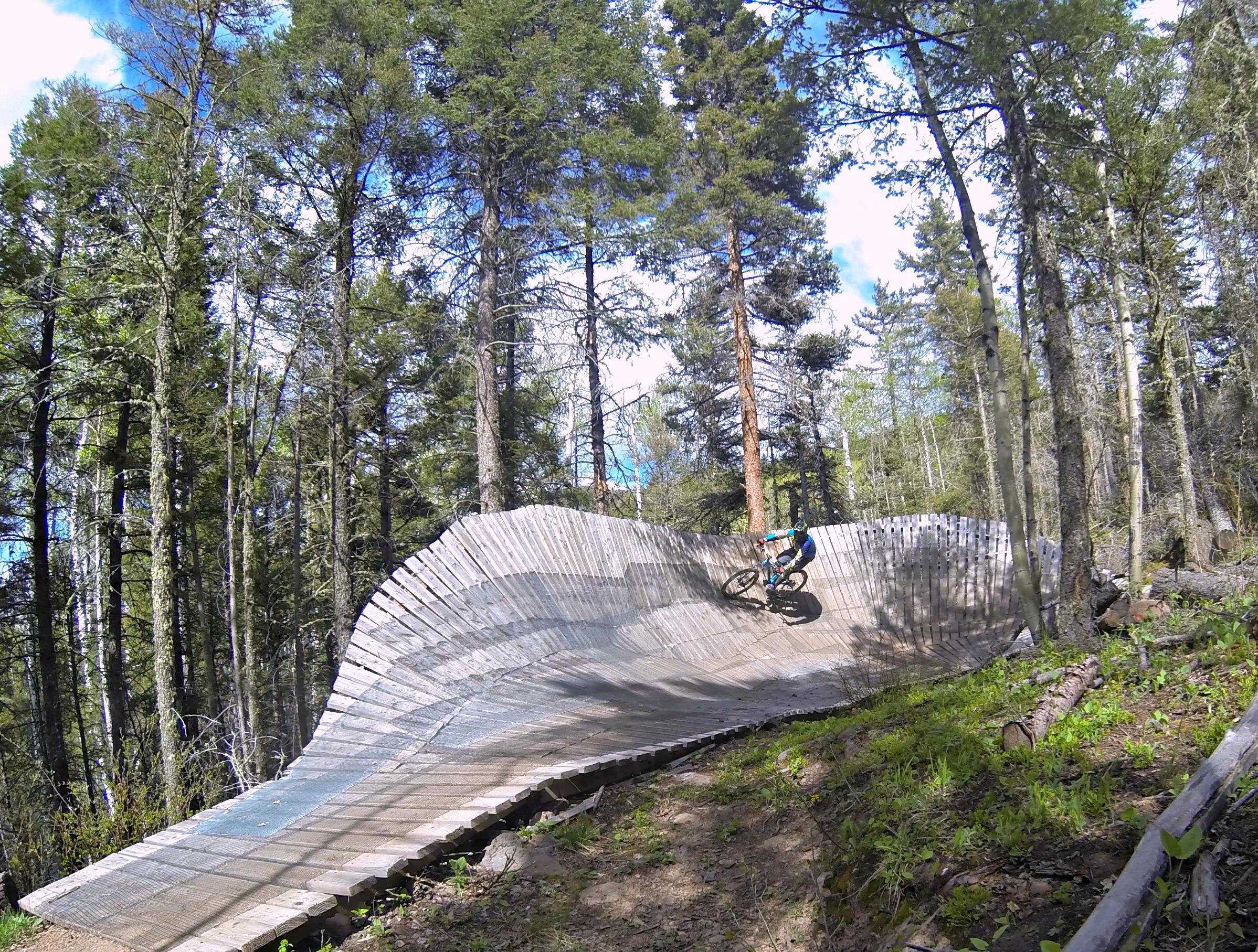 A mountain biker rides a wooden pump track surrounded by tall trees in a forest. The track features a curved wooden structure, allowing for an exhilarating downhill ride, with a bright blue sky peeking through the foliage above. Angel Fire Bike Park mountain bike trail.