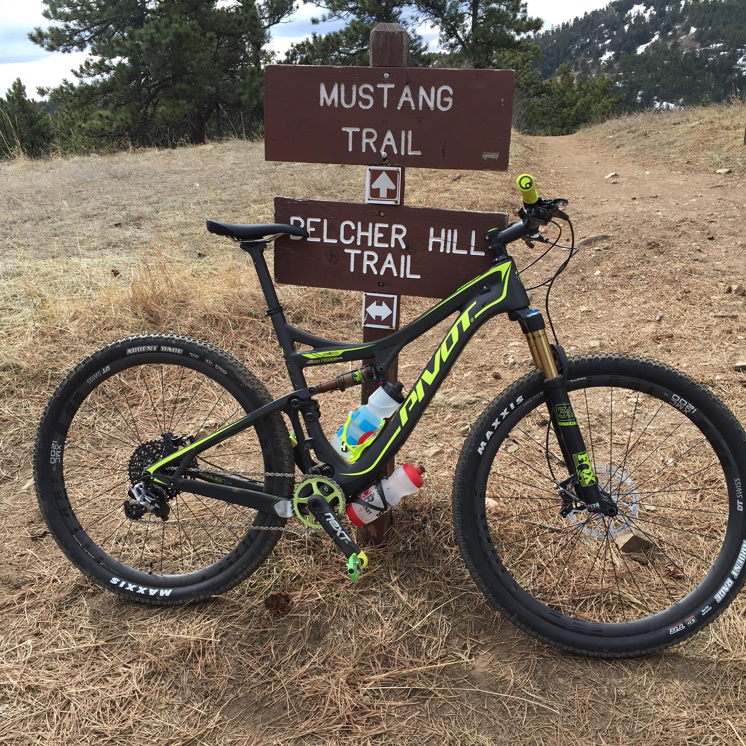 Pivot Mach 429SL Carbon: A mountain bike parked next to a trail sign indicating the Mustang Trail and Belcher Hill Trail, surrounded by grassy terrain and pine trees, with a backdrop of hills and mountains.