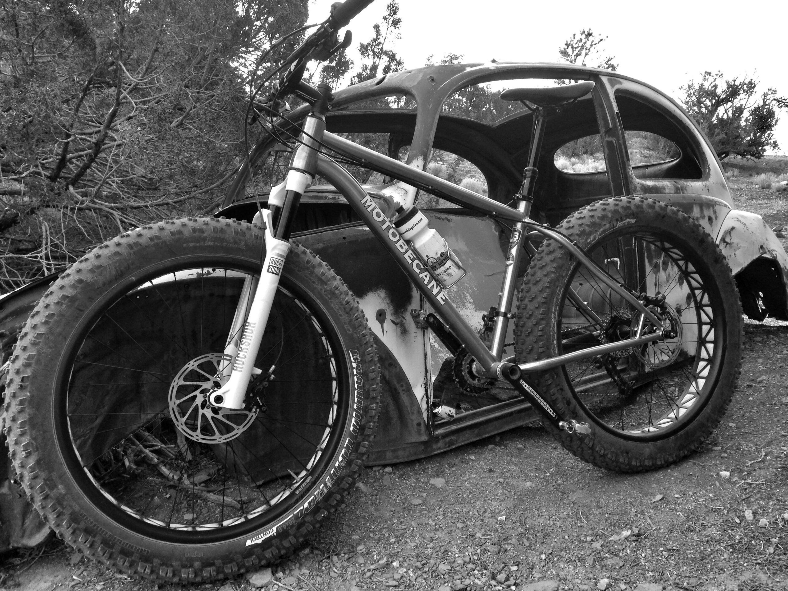 A black and white photograph of a mountain bike resting against the side of an old, rusted car. The bike features large, knobby tires and a metal frame, with a water bottle attached to the frame. In the background, there are trees and overgrown vegetation, adding to the rustic atmosphere of the scene. Buggy mountain bike trail.