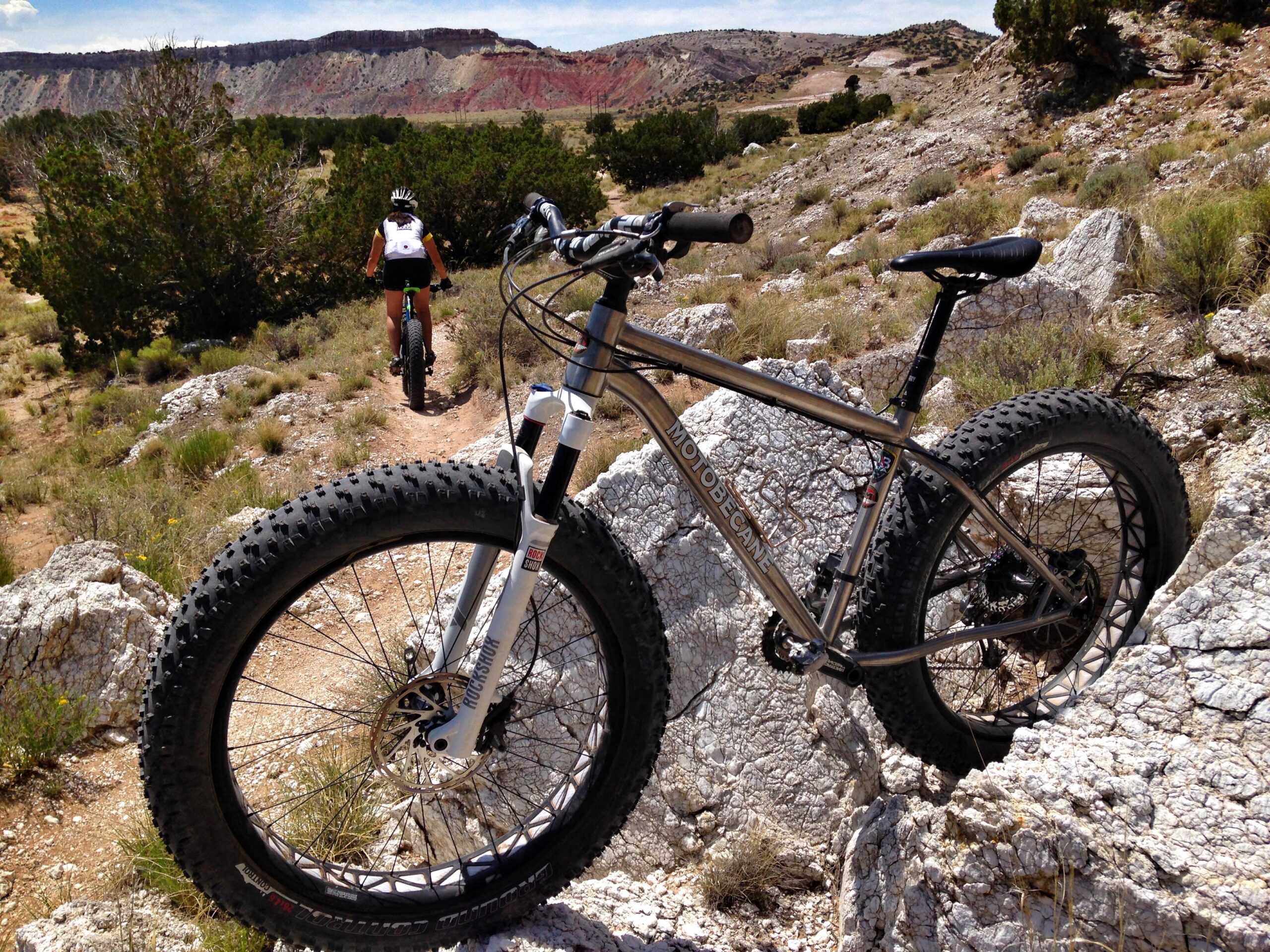 A close-up of a fat-tire mountain bike resting on rocky terrain, with a trail and a mountainous landscape visible in the background. A cyclist is riding away along the path, surrounded by greenery and rugged hills under a clear blue sky. The bike features wide tires designed for off-road riding. White Ridge Bike Trails mountain bike trail.