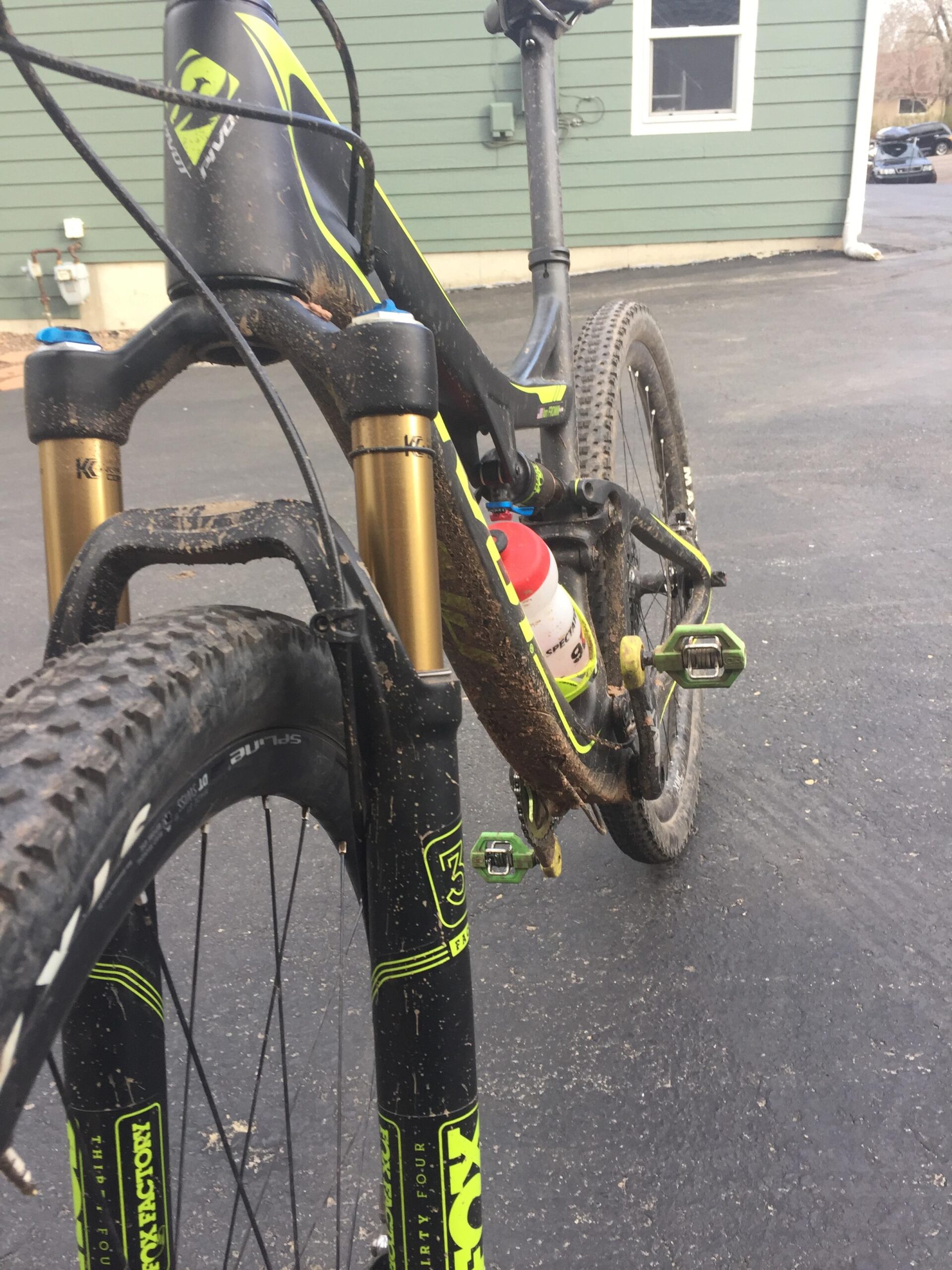 Pivot Mach 429SL Carbon: A close-up view of a mountain bike, showcasing its front fork and frame, which are covered in mud, suggesting recent outdoor use. A water bottle is mounted in the frame. The background features a green house exterior and a paved surface.