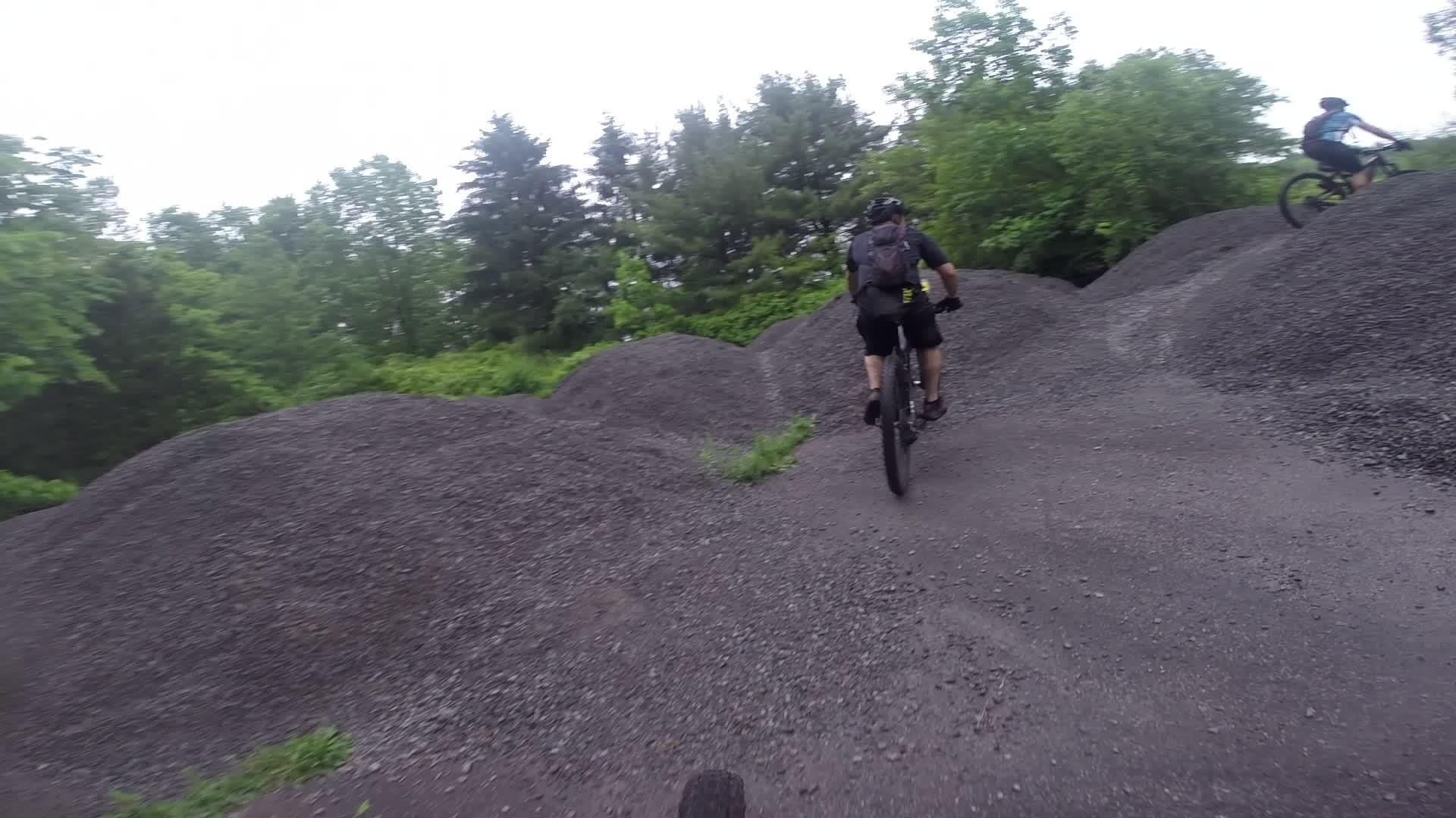 A group of mountain bikers riding on a gravel path, surrounded by large piles of gravel and lush greenery. The scene is set under a cloudy sky, showcasing an adventurous biking trail. Stewart State Forest mountain bike trail.