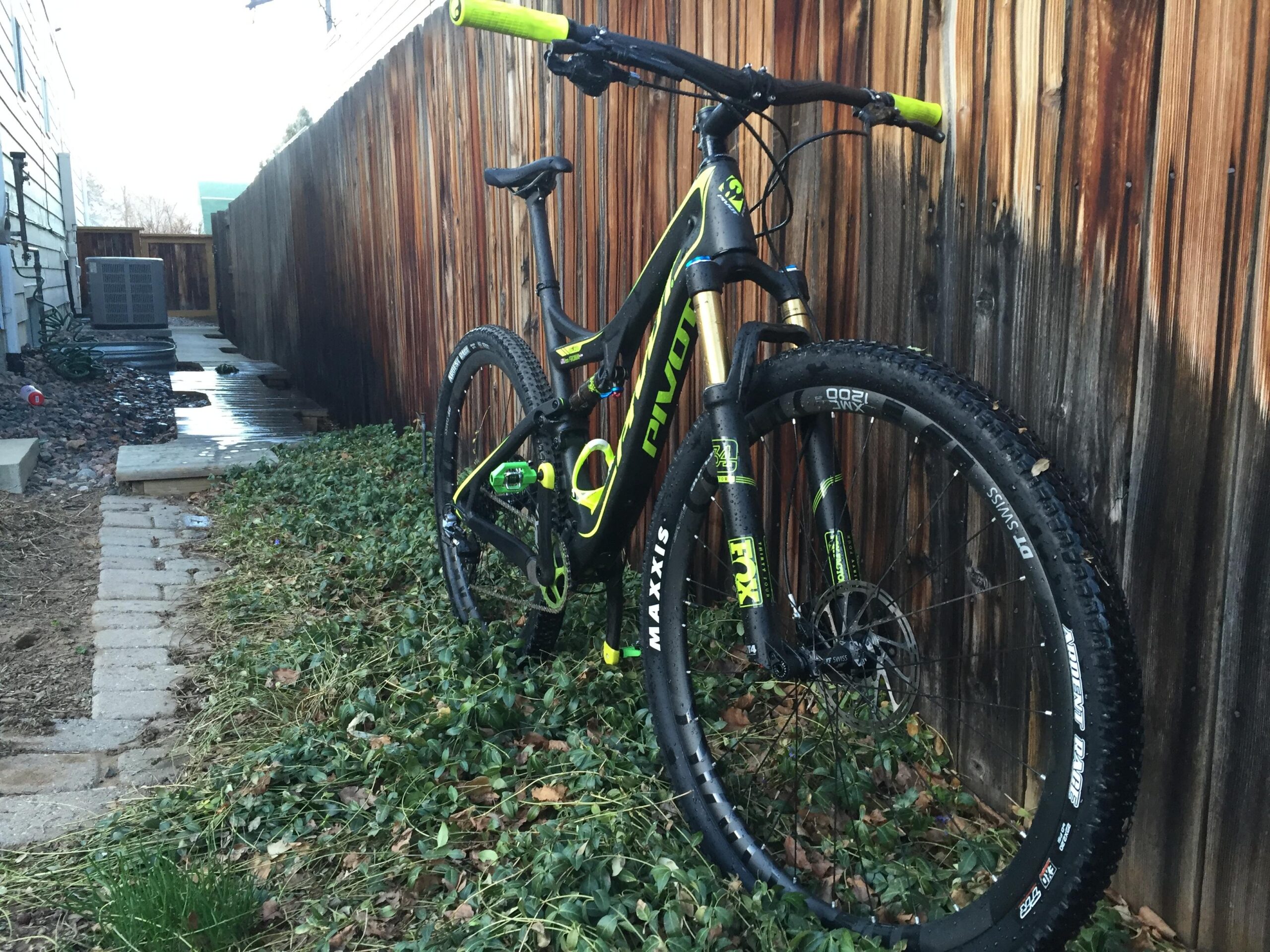 Pivot Mach 429SL Carbon: A black and yellow mountain bike parked beside a wooden fence, with a narrow walkway covered in gravel and weeds. The bike features distinctive branding and thick tires, indicating its suitability for off-road riding.