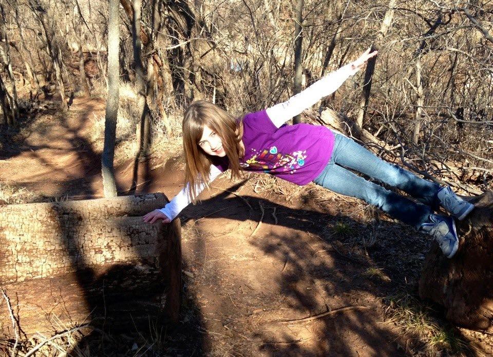 A child playfully balances on a log in a wooded area, extending her arms out to the sides and smiling. She is wearing a purple shirt and blue jeans, surrounded by trees and earthy ground. Bluff Creek Trail mountain bike trail.