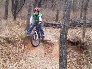 A young boy wearing a helmet and a green shirt is riding a bicycle on a dirt trail in a wooded area, balancing while navigating a small jump over a dip in the terrain. Leaves cover the ground, and trees are visible in the background. Thunderbird Lake Clear Bay mountain bike trail.
