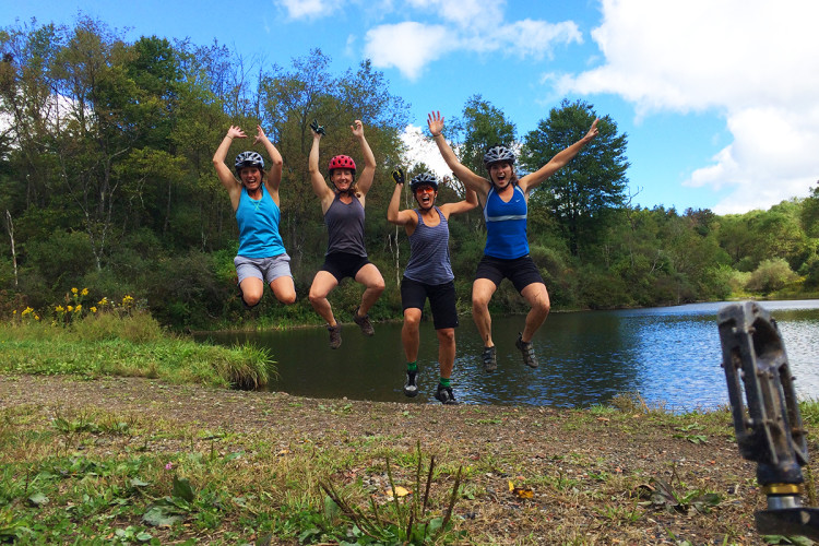Four women wearing helmets and active wear are joyfully jumping in front of a lake surrounded by greenery. They are all smiling and have their arms raised in excitement. The sky is partly cloudy, adding a vibrant atmosphere to the scene. A bicycle pedal is visible in the foreground.