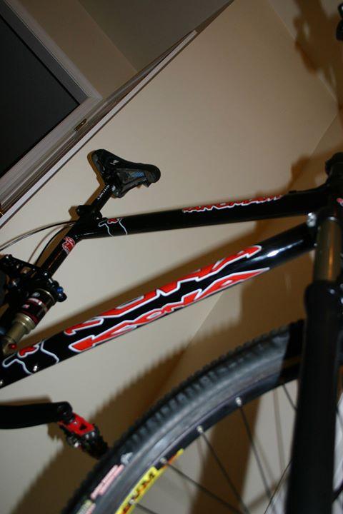 Kona ManoMano: A black mountain bike frame is photographed from a low angle, showcasing its design and components against a neutral wall background. The bike features prominent red and white logos, with visible gears and a thick tire in focus.