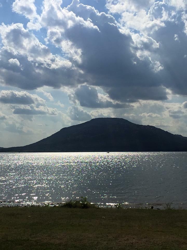 A scenic view of a lake with shimmering water reflecting sunlight, framed by a distant mountain under a partly cloudy sky. The shoreline features patches of grass and vegetation in the foreground. Lake Lawtonka Trails mountain bike trail.