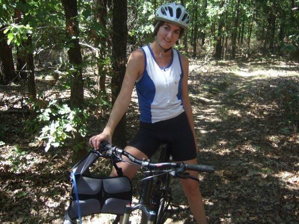 A young woman wearing a helmet and athletic cycling gear stands beside her mountain bike on a wooded trail, smiling at the camera. The background features trees and a dirt path, indicating an outdoor biking environment. McMurtry Trail mountain bike trail.