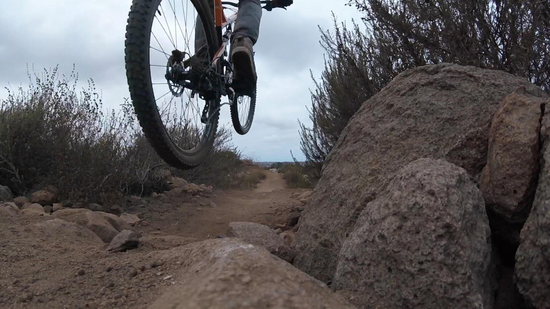 Haro Shift R5: A mountain biker jumps over a rocky path, showcasing the bike's rear wheel elevated off the ground. Surrounding the trail are patches of shrubs and vegetation under a cloudy sky. The image captures the action and excitement of off-road cycling.