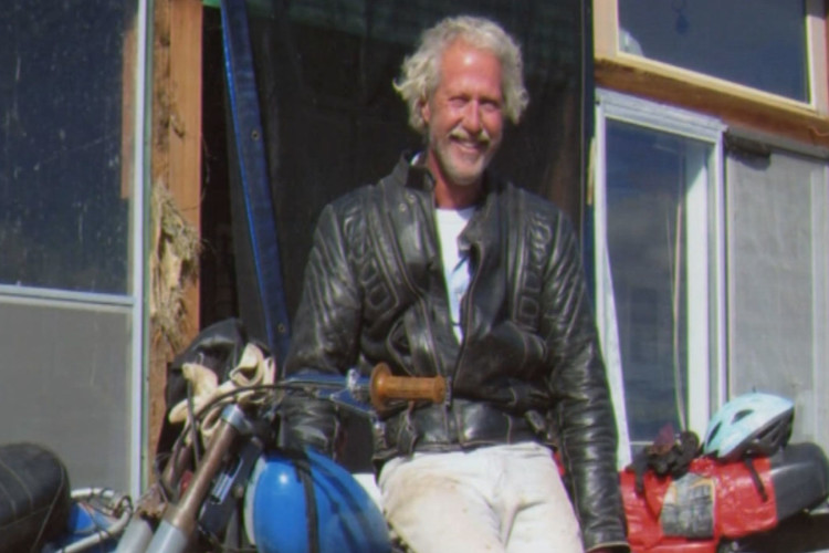 A smiling man with curly hair, wearing a black leather motorcycle jacket and white pants, sits on a motorcycle in front of a building with large windows. Various helmets and motorcycle gear are visible in the background. The scene has a bright, sunny ambiance.